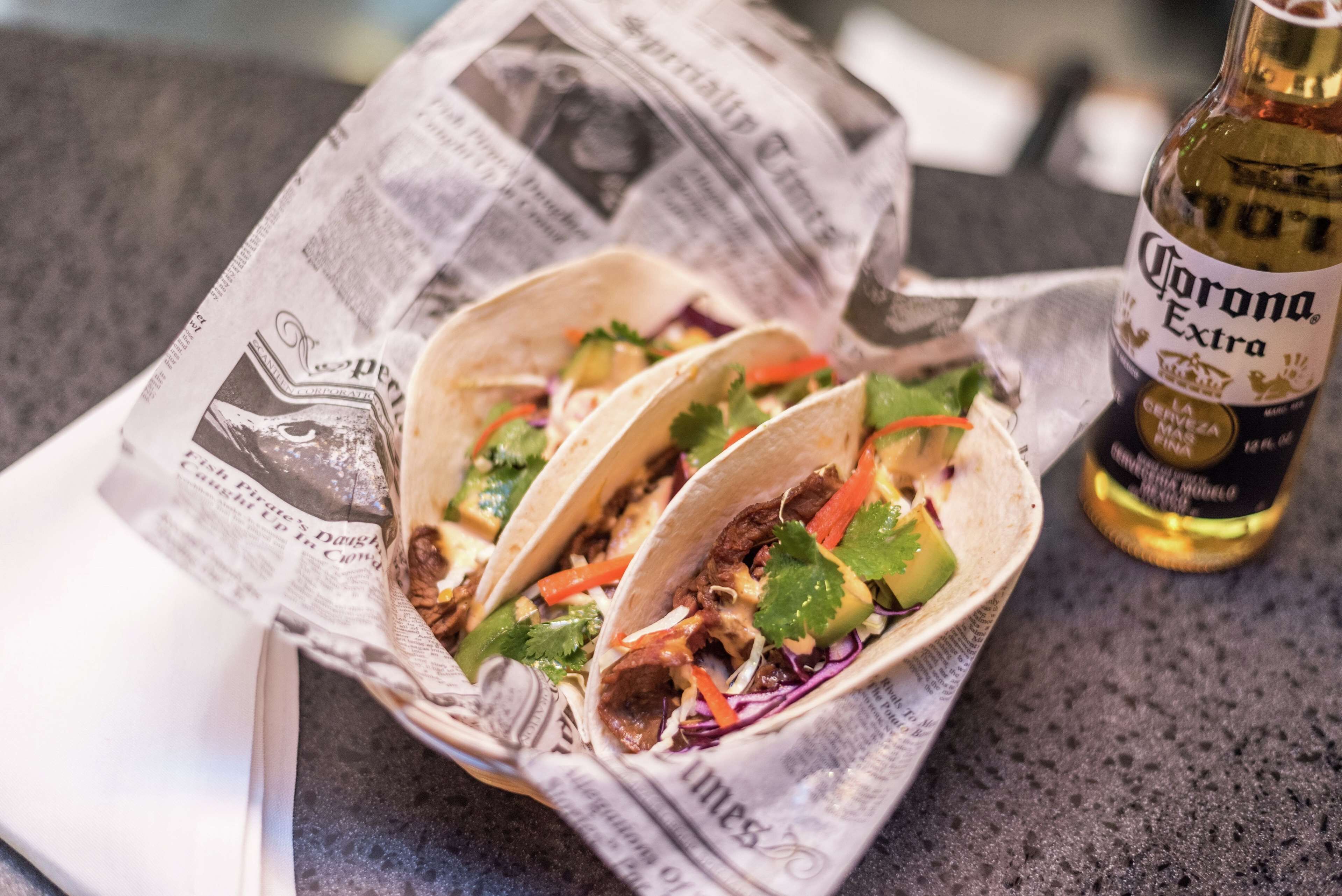 Trois tacos sur un journal, avec une bouteille de bière Corona Extra à proximité.