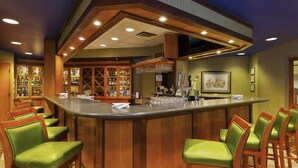A bar area with a wooden bar counter, green cushioned bar stools, and a wine rack with bottles.