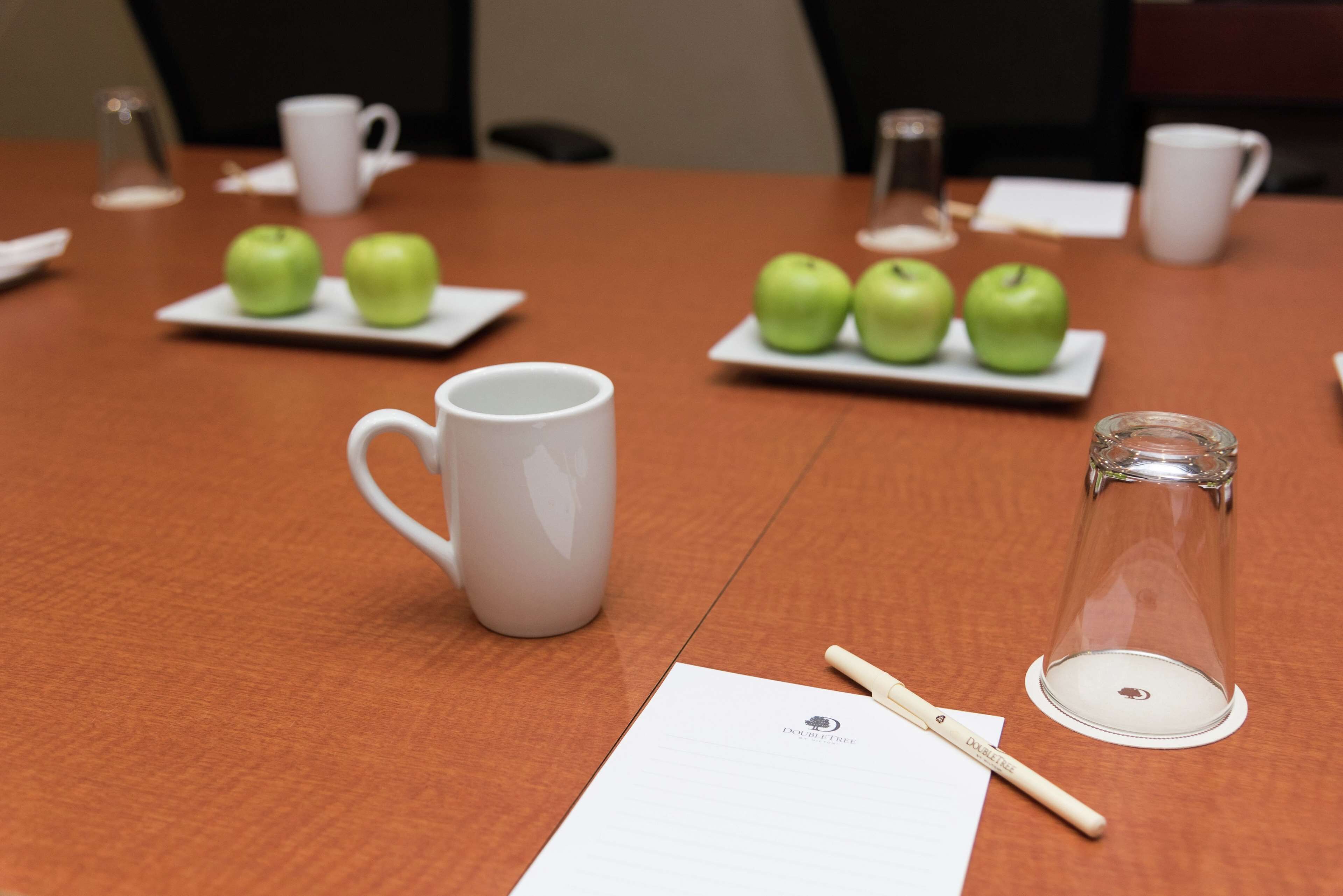 A conference table with a white mug, a glass, a pen, and two plates with green apples.