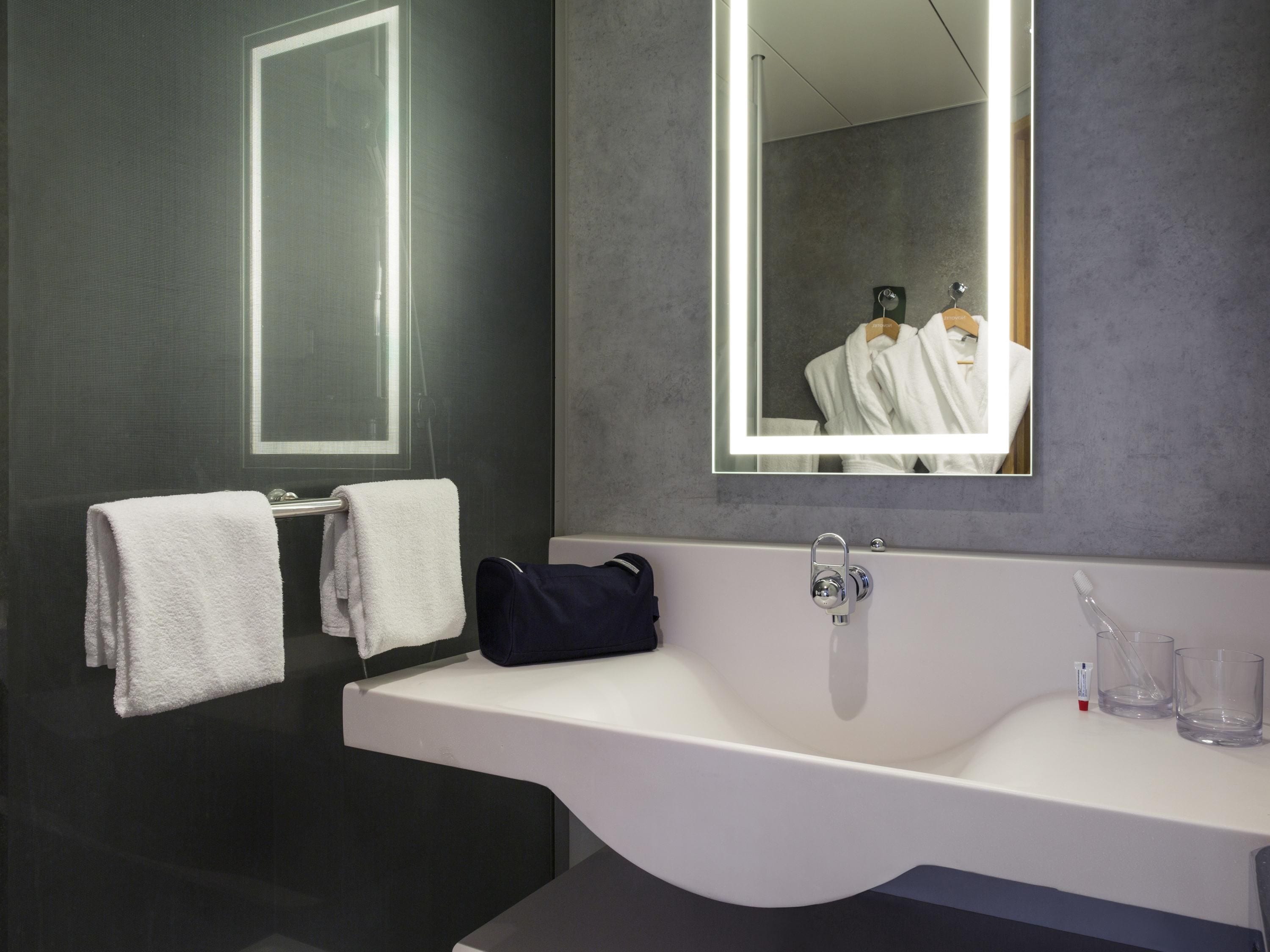 A modern bathroom with a large mirror, two white towels, a white sink, and a dark-colored bag on the counter.