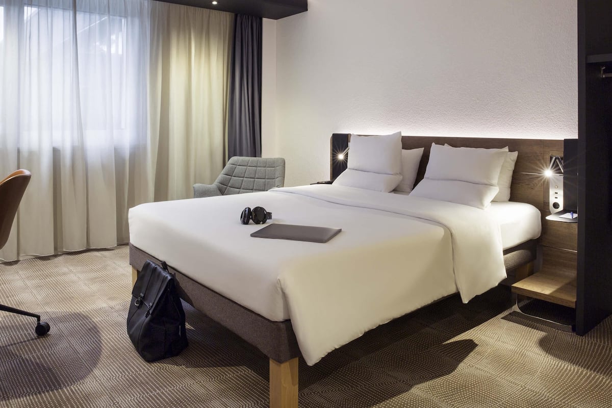 A hotel room with a large bed, a chair, a desk with a lamp, and a window with curtains.