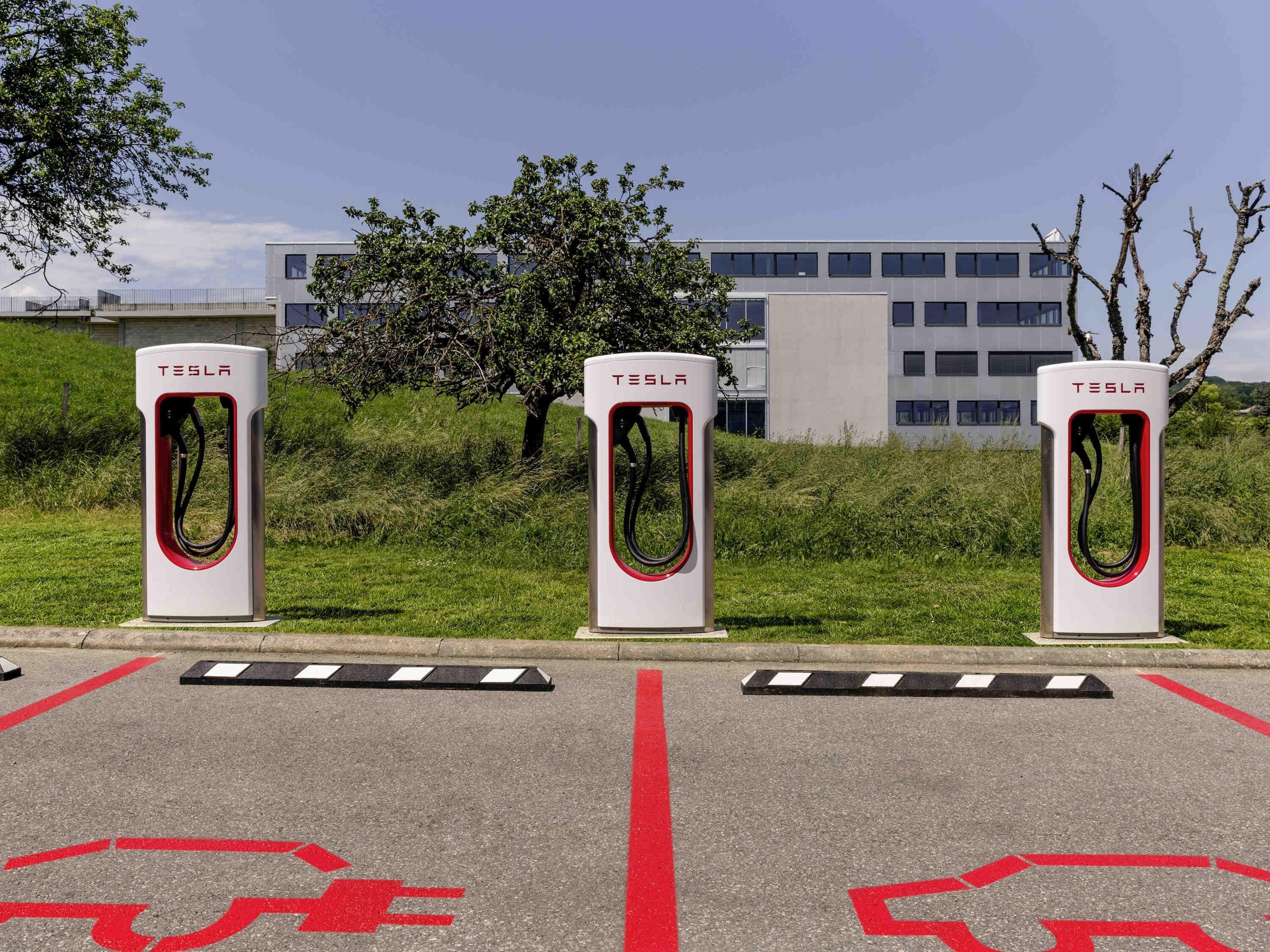 Three Tesla electric vehicle charging stations in a parking lot with a building in the background.