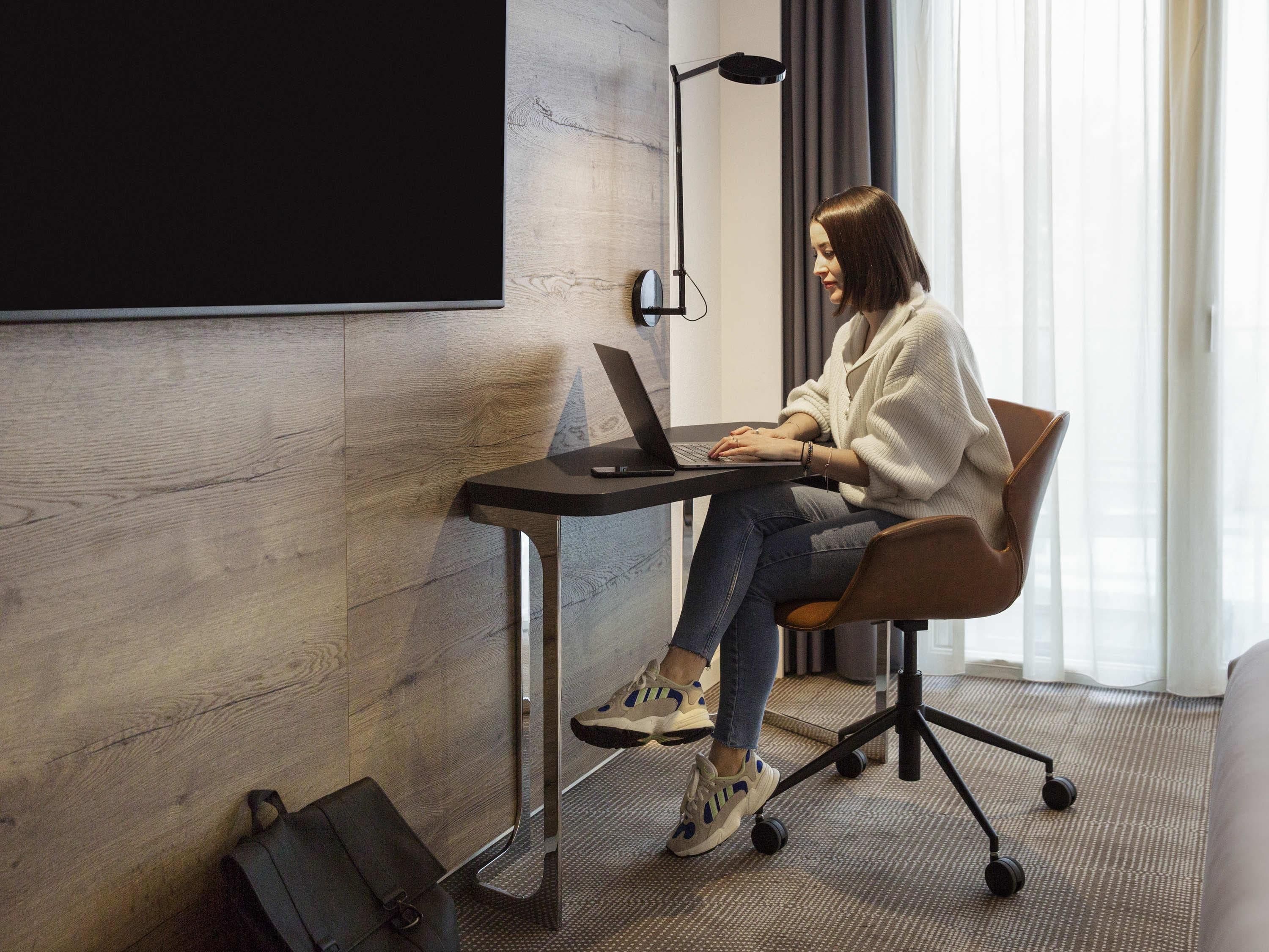 Eine Person sitzt an einem Schreibtisch mit einem Laptop in einem Raum mit Holzwand und Projektionsschirm.