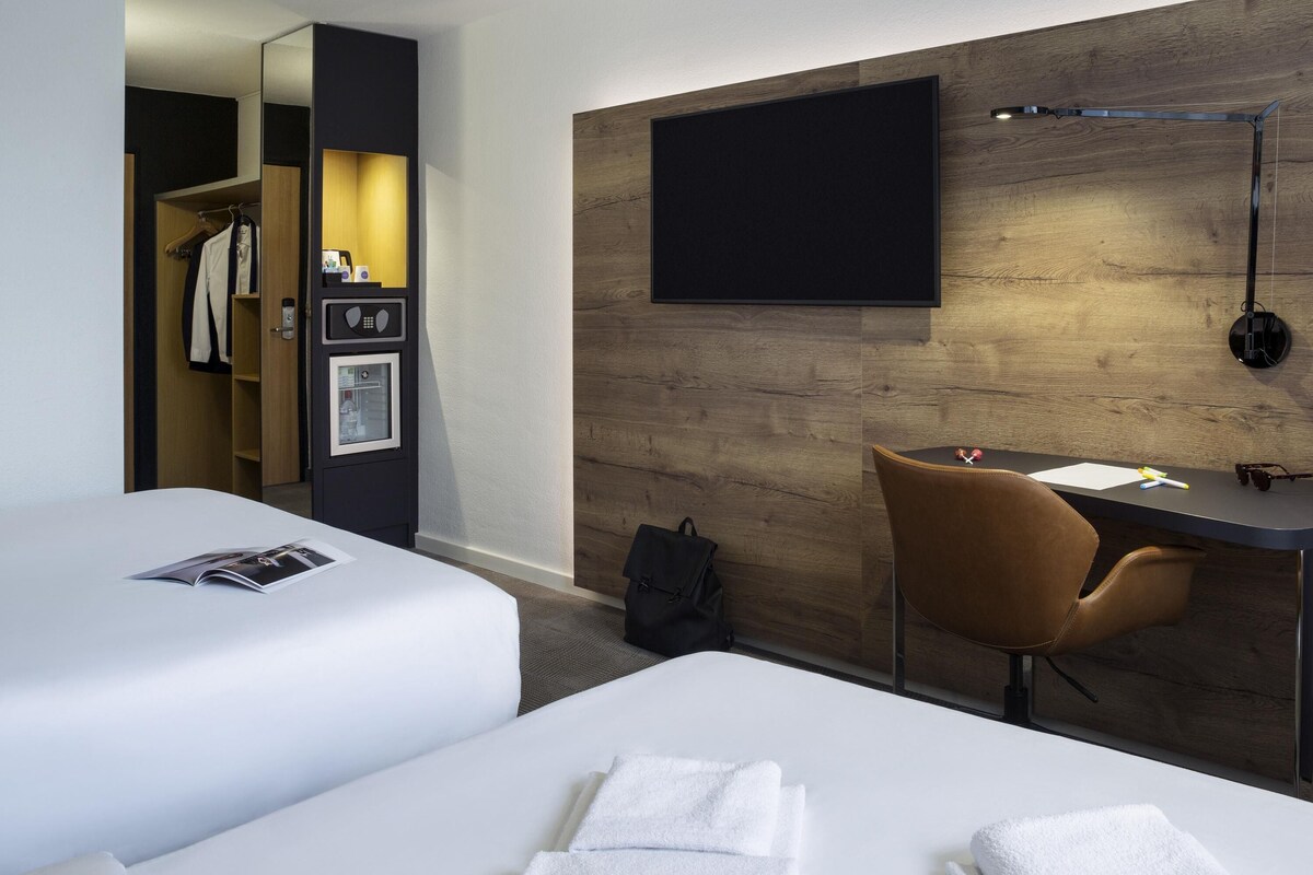 A hotel room with two beds, a desk, a chair, a wardrobe, and a TV mounted on the wall.