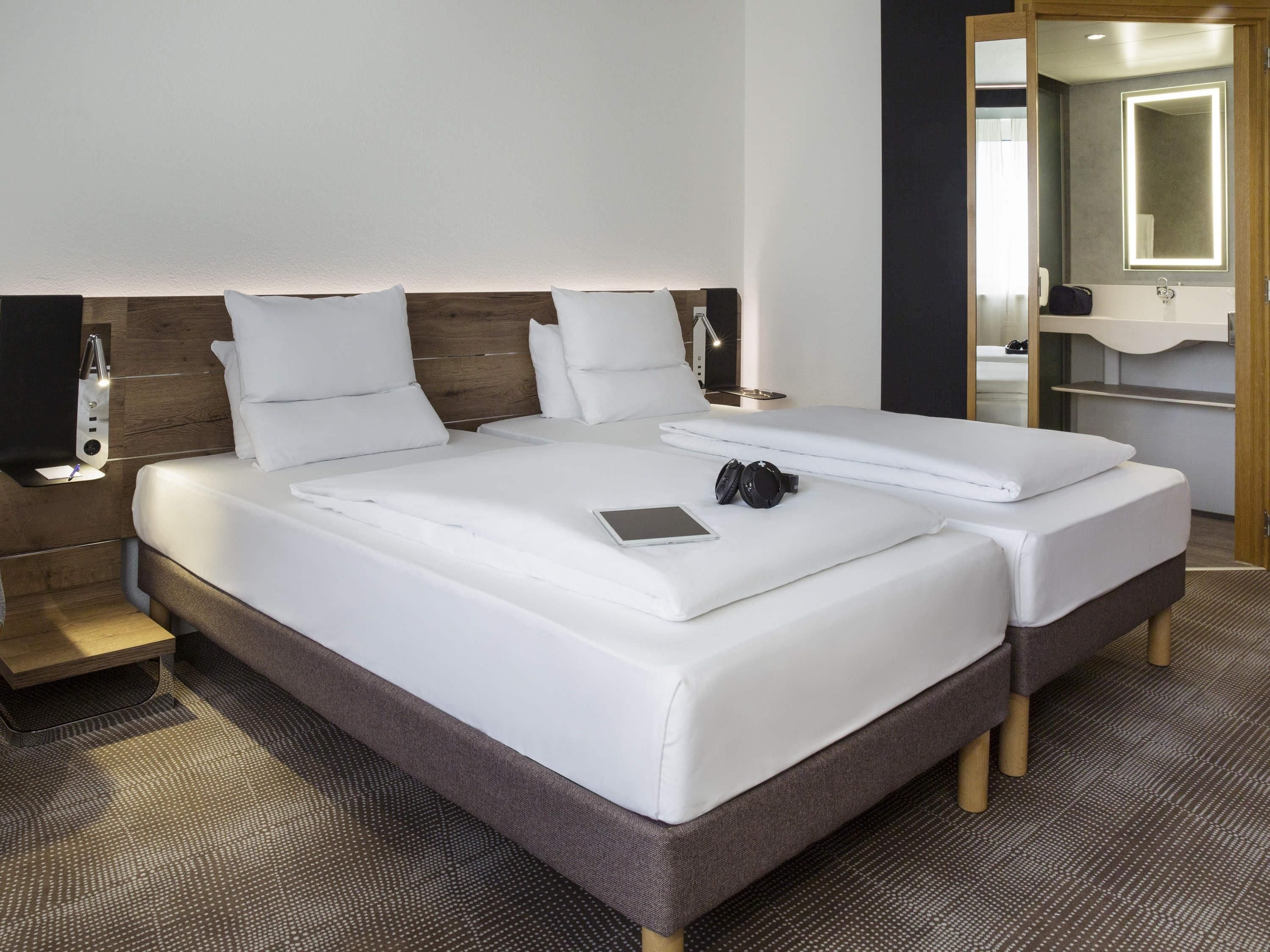 Cama doble con sábanas blancas, mesita de noche con lámpara y espejo sobre el lavabo.