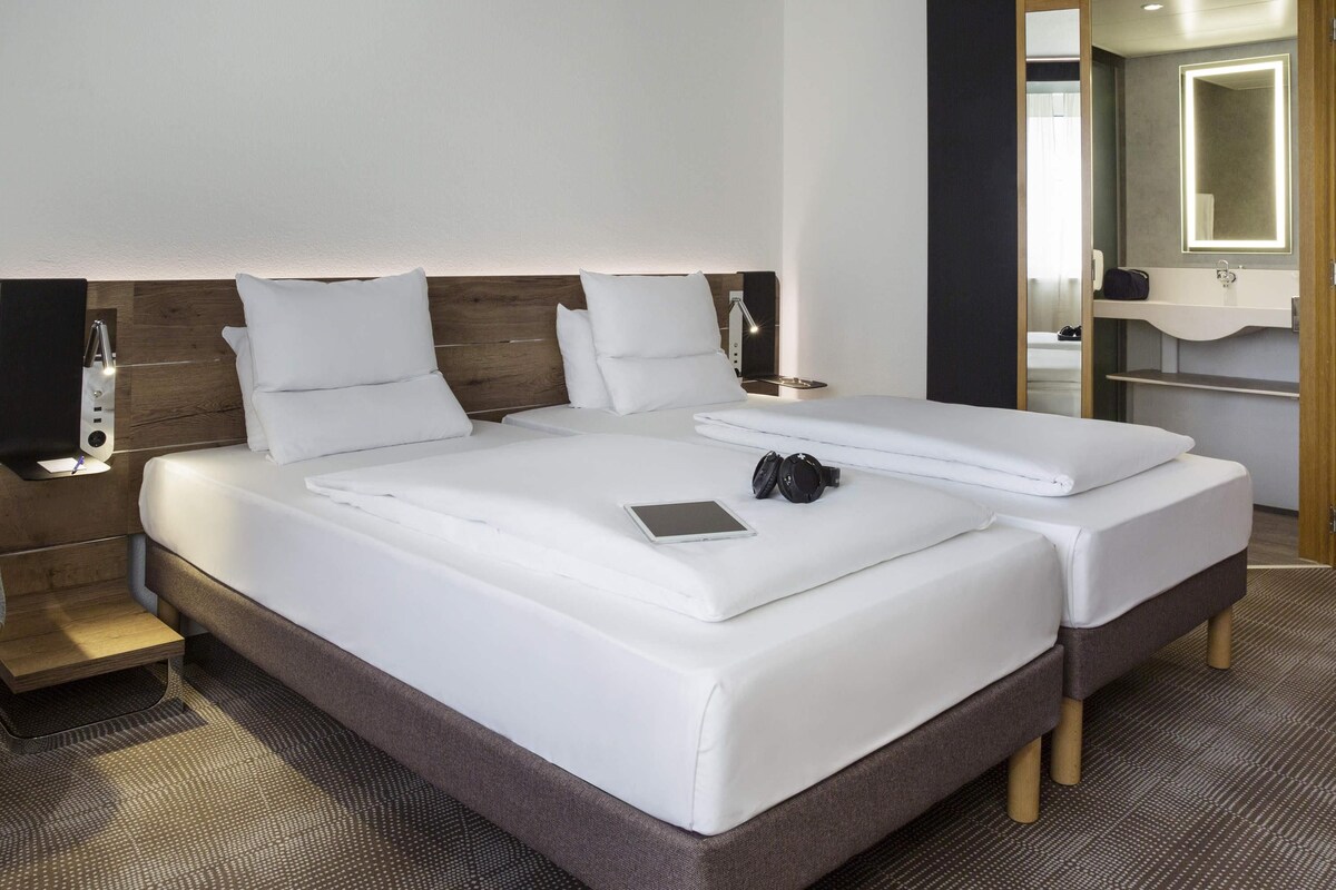 A double bed with white linens, a bedside table with a lamp, and a mirror above the sink.
