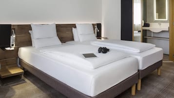 Cama doble con sábanas blancas, mesita de noche con lámpara y espejo sobre el lavabo.