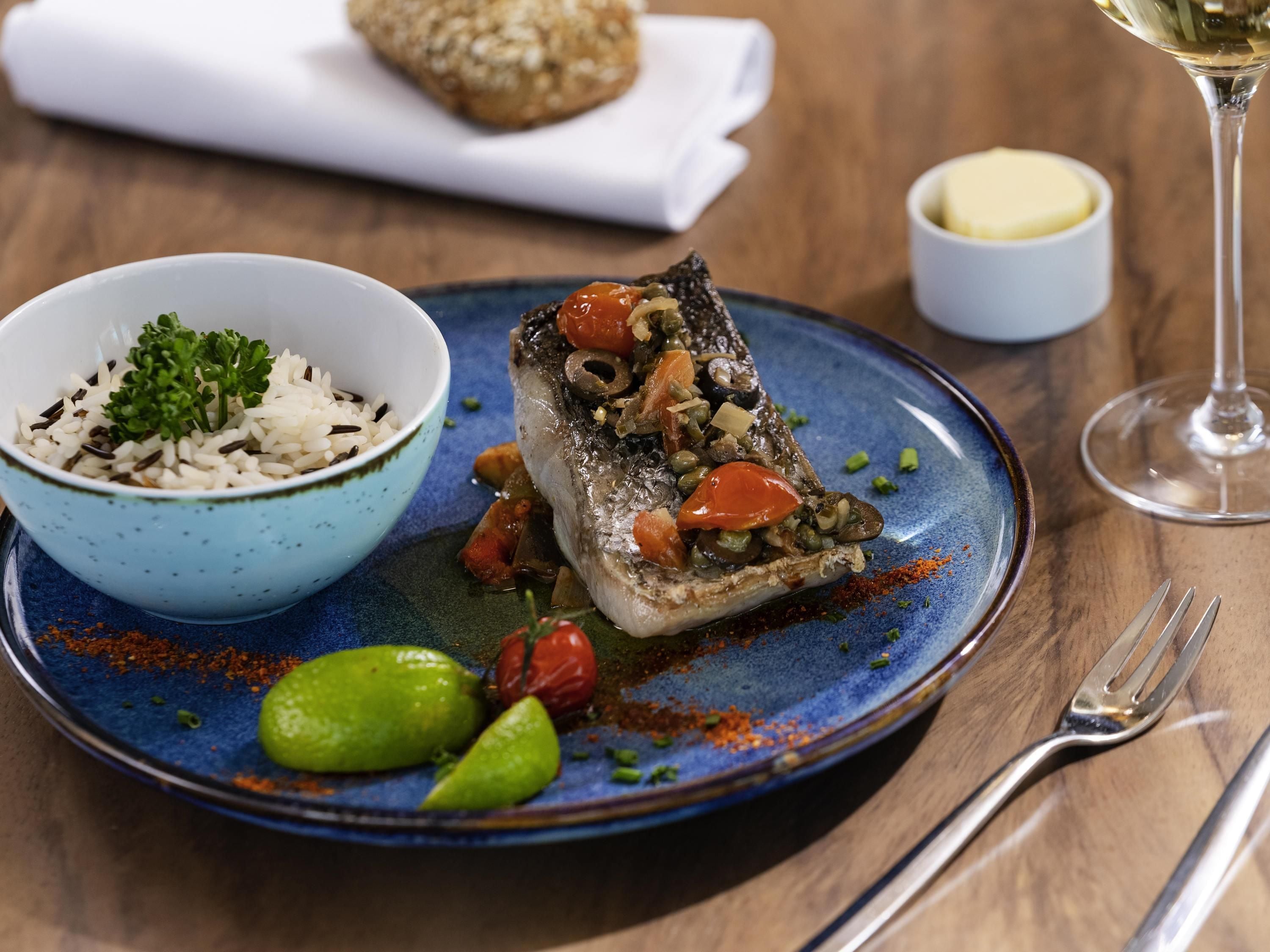 Une assiette contenant un plat de poisson, un bol de riz et un verre de vin blanc, le tout posé sur une table en bois.
