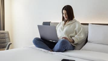 Une personne assise sur un lit, avec un ordinateur portable, une télécommande et un oreiller.