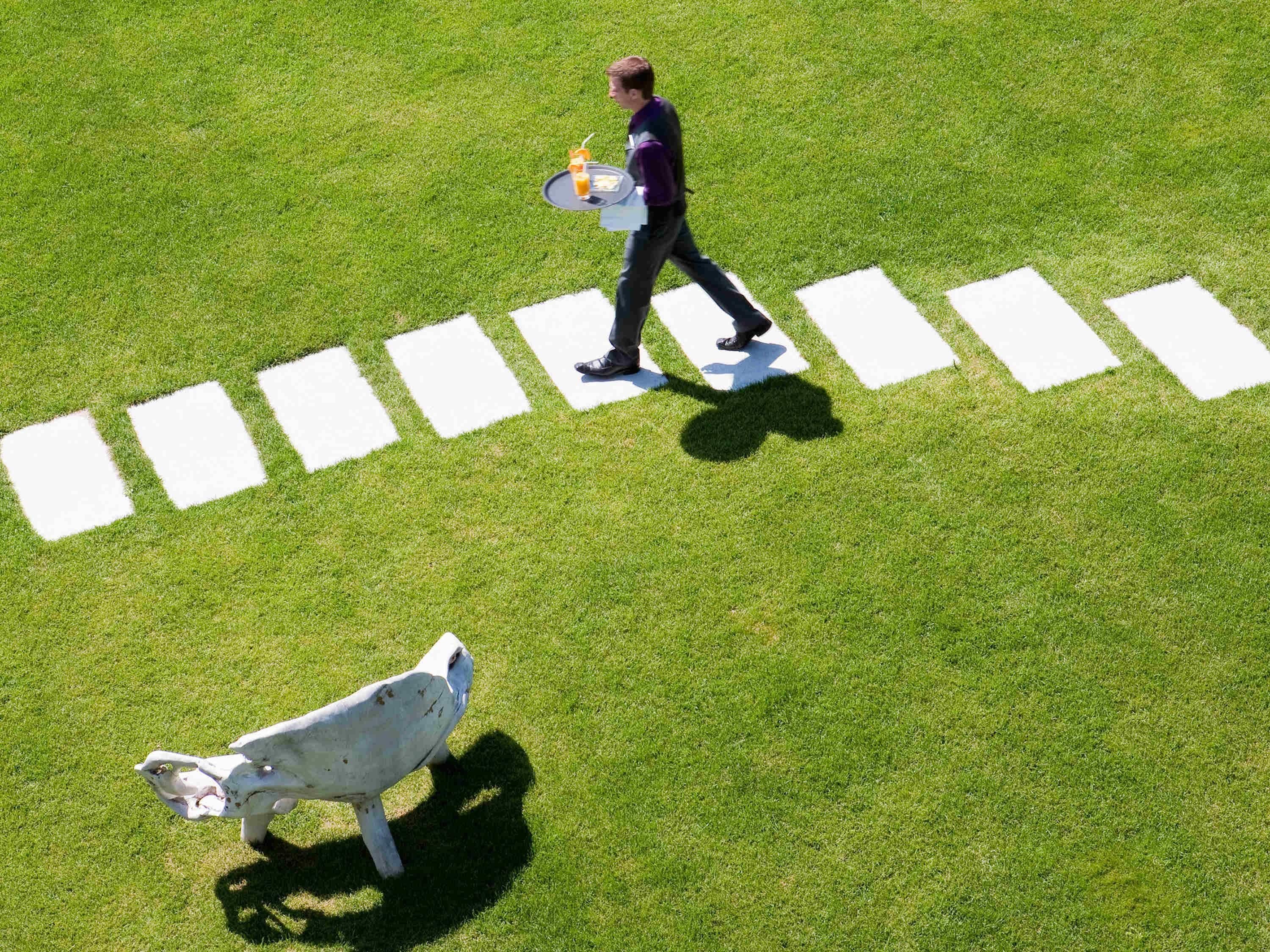 A person walking across a grassy field with white markings, carrying a tray with a drink and food.