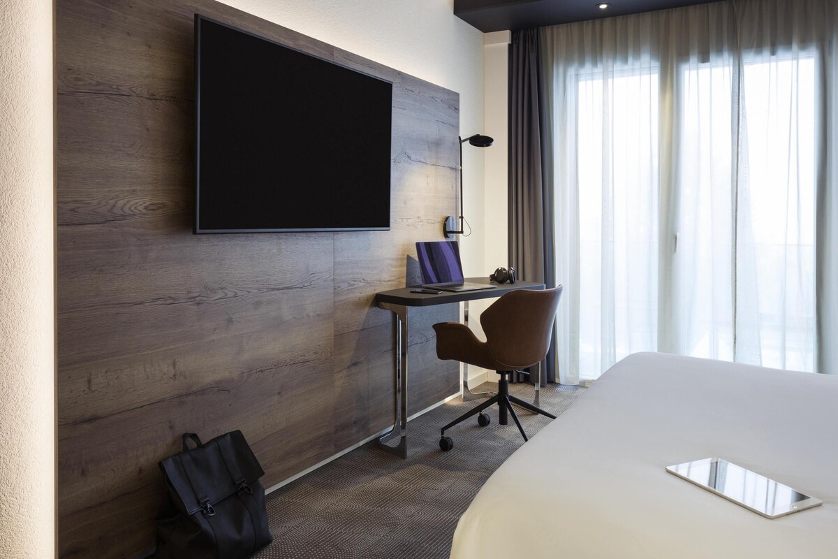 A hotel room with a large bed, a desk with a chair, a television mounted on the wall, and a window with curtains.