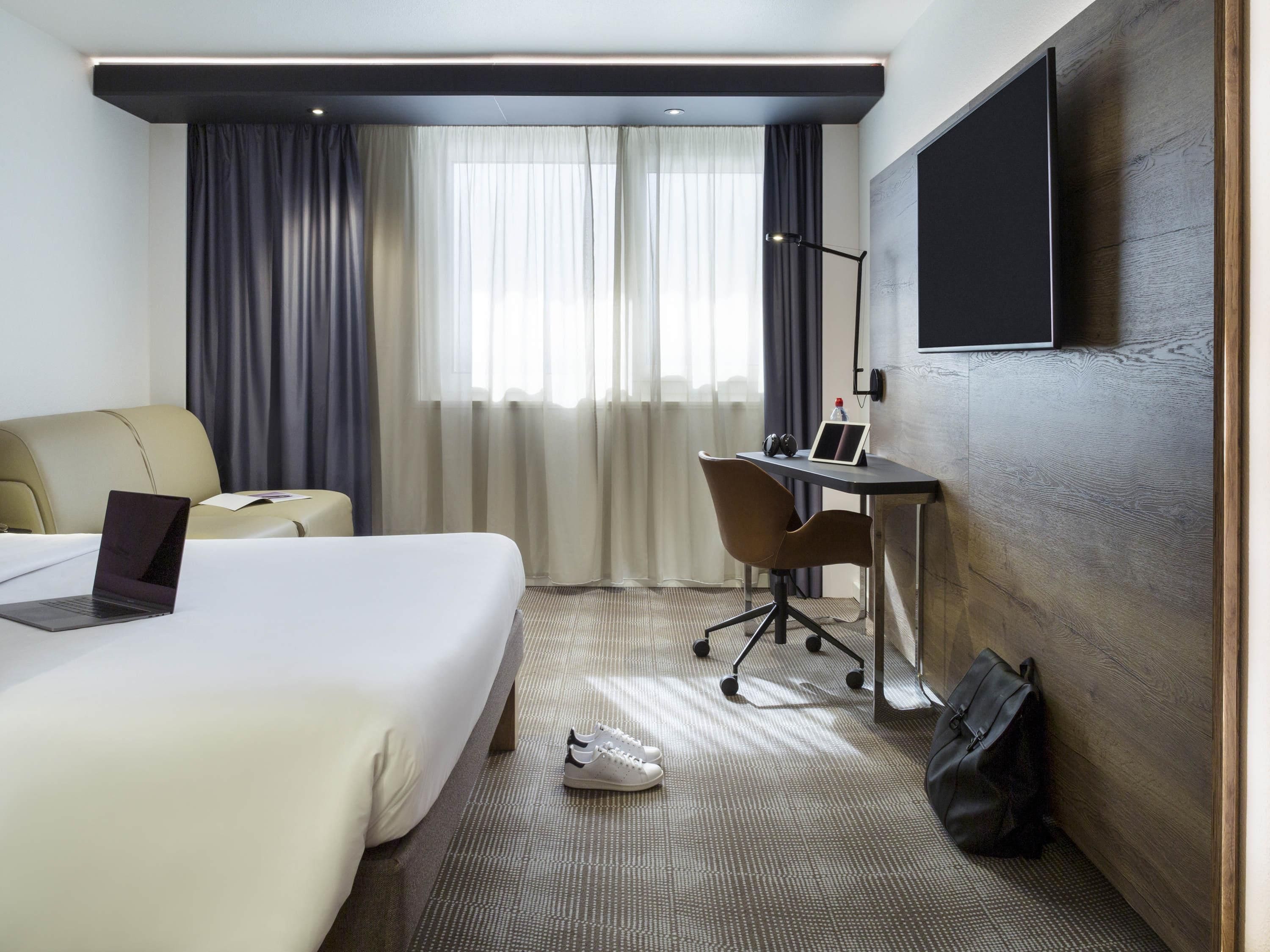 A hotel room with a bed, a desk, a chair, a laptop, a TV, and a bag.