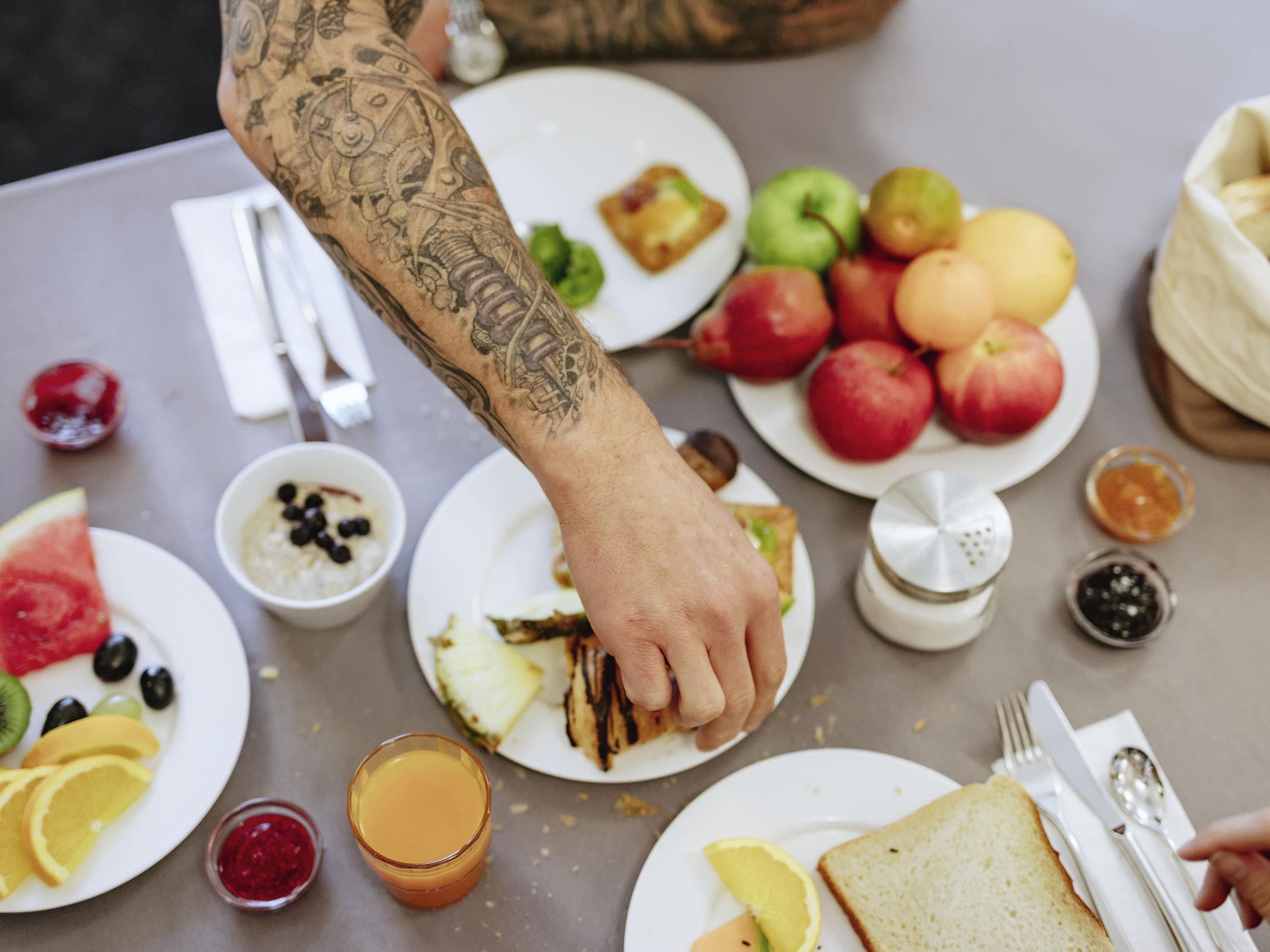 A dining table with plates of food, a glass of orange juice, and a person's arm with a tattoo.