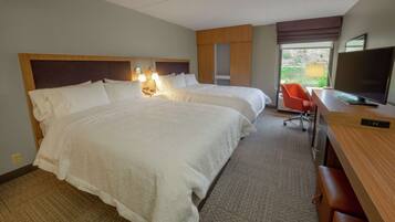 A hotel room with two beds, a desk with a TV, a chair, and a window with a view of greenery.