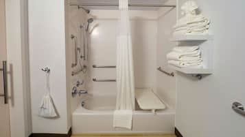 A modern bathroom with a bathtub, shower, and toilet. There are grab bars, a towel rack, and a shelf with folded towels.