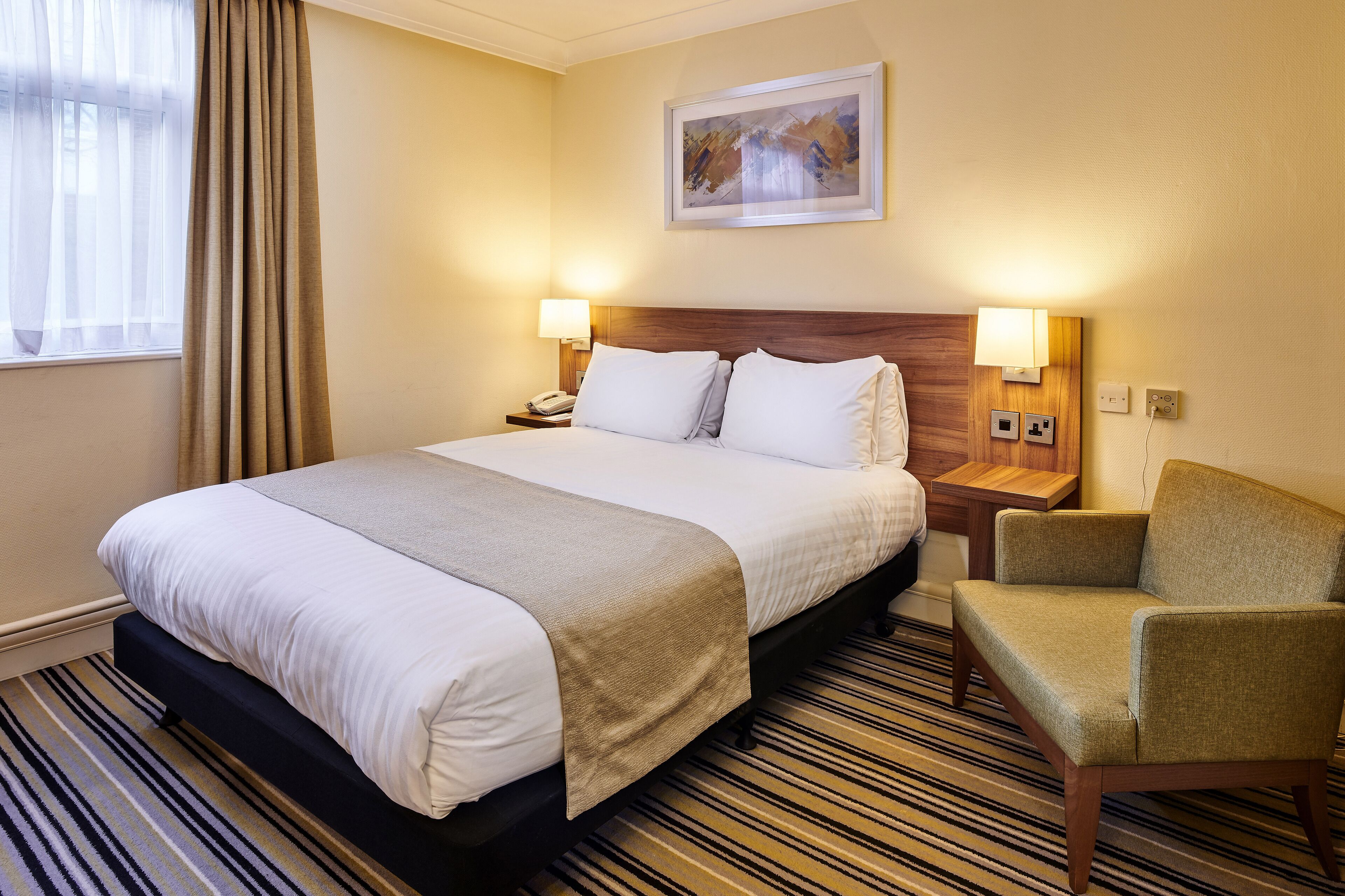 A hotel room with a large bed, two bedside lamps, a chair, a painting on the wall, and a striped carpet.