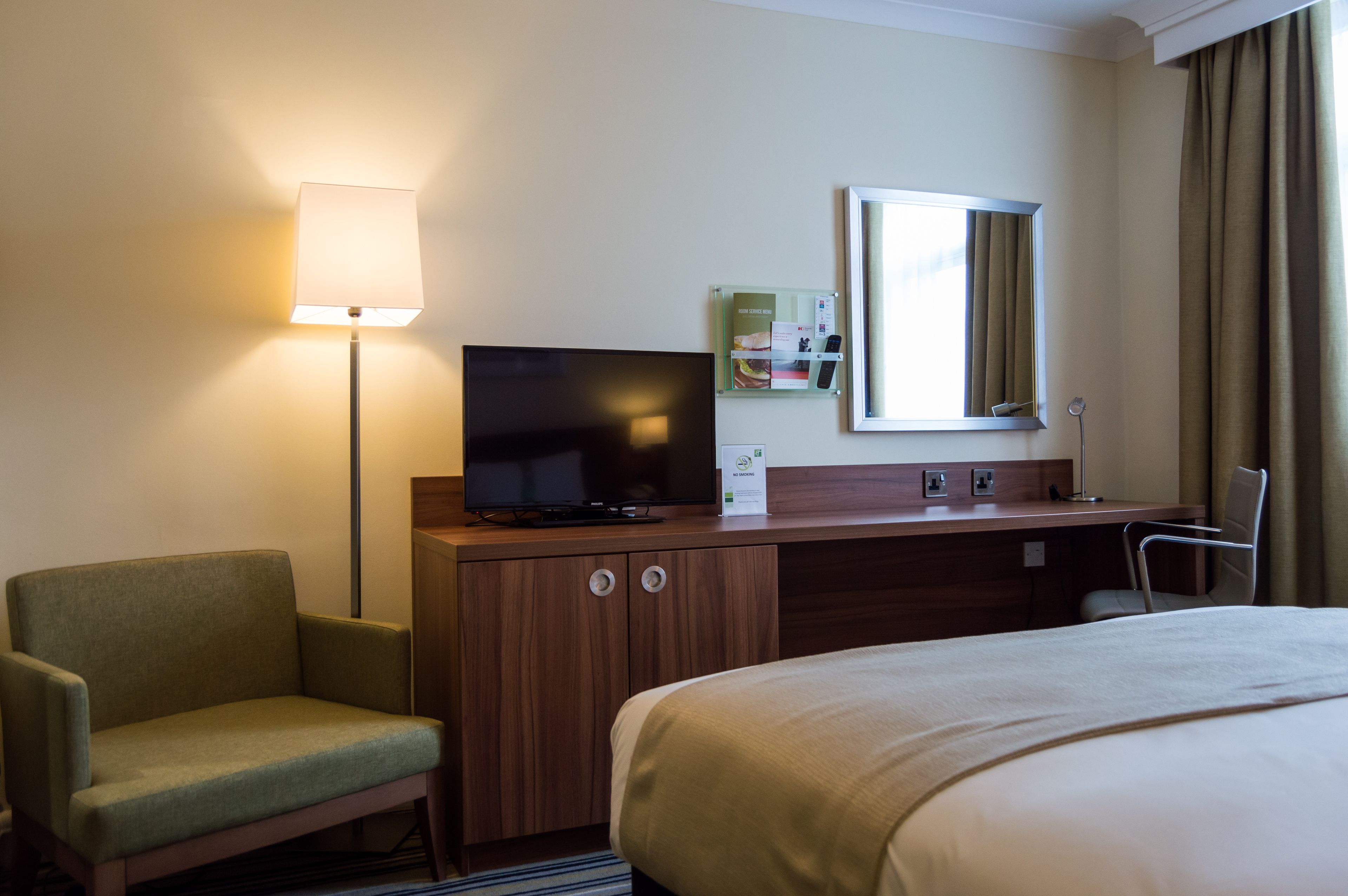 A hotel room with a bed, a desk with a television, a chair, a lamp, and a mirror.