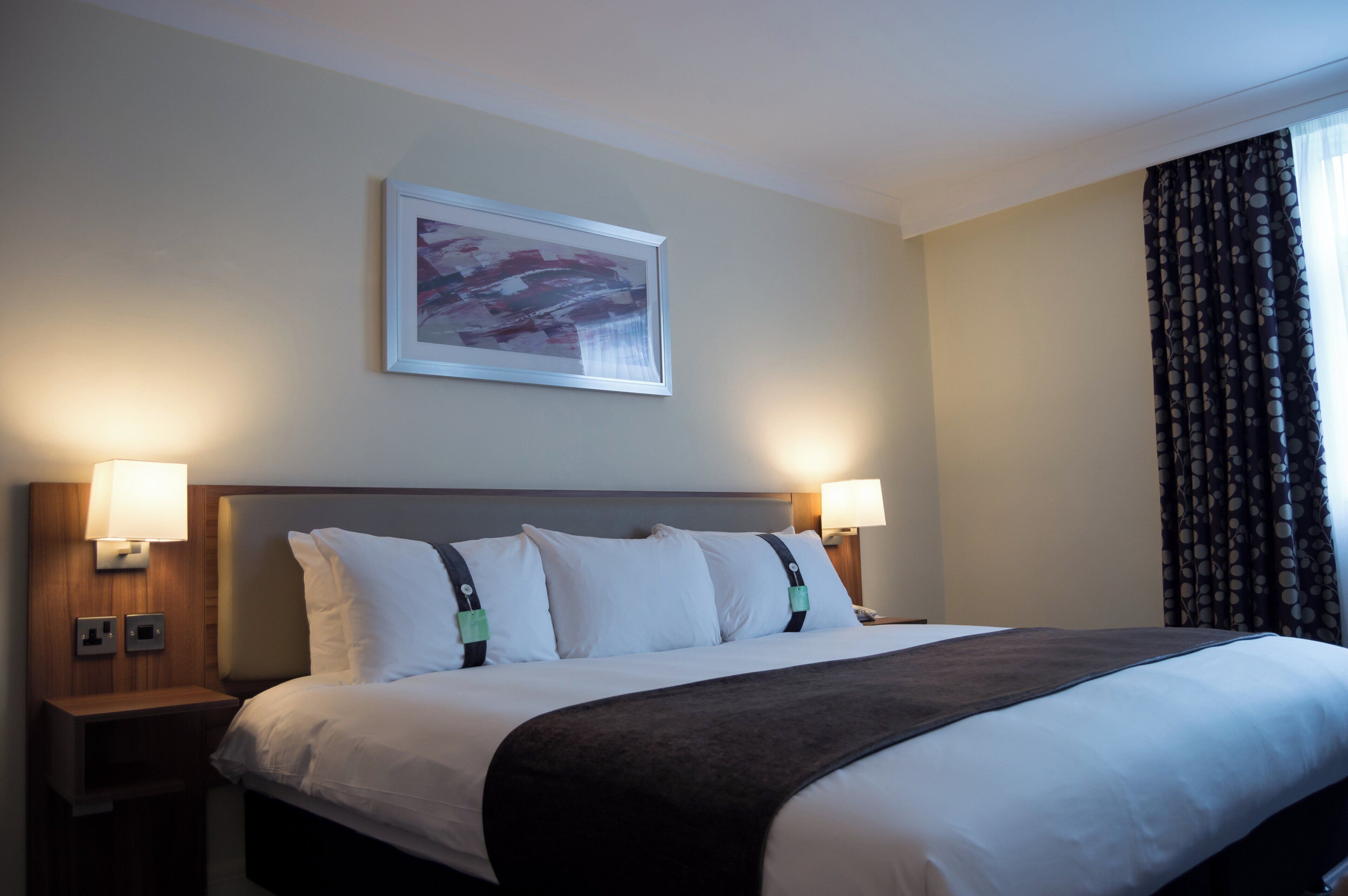 A hotel room with a bed, two bedside lamps, a nightstand, a wall-mounted picture, and a window with curtains.