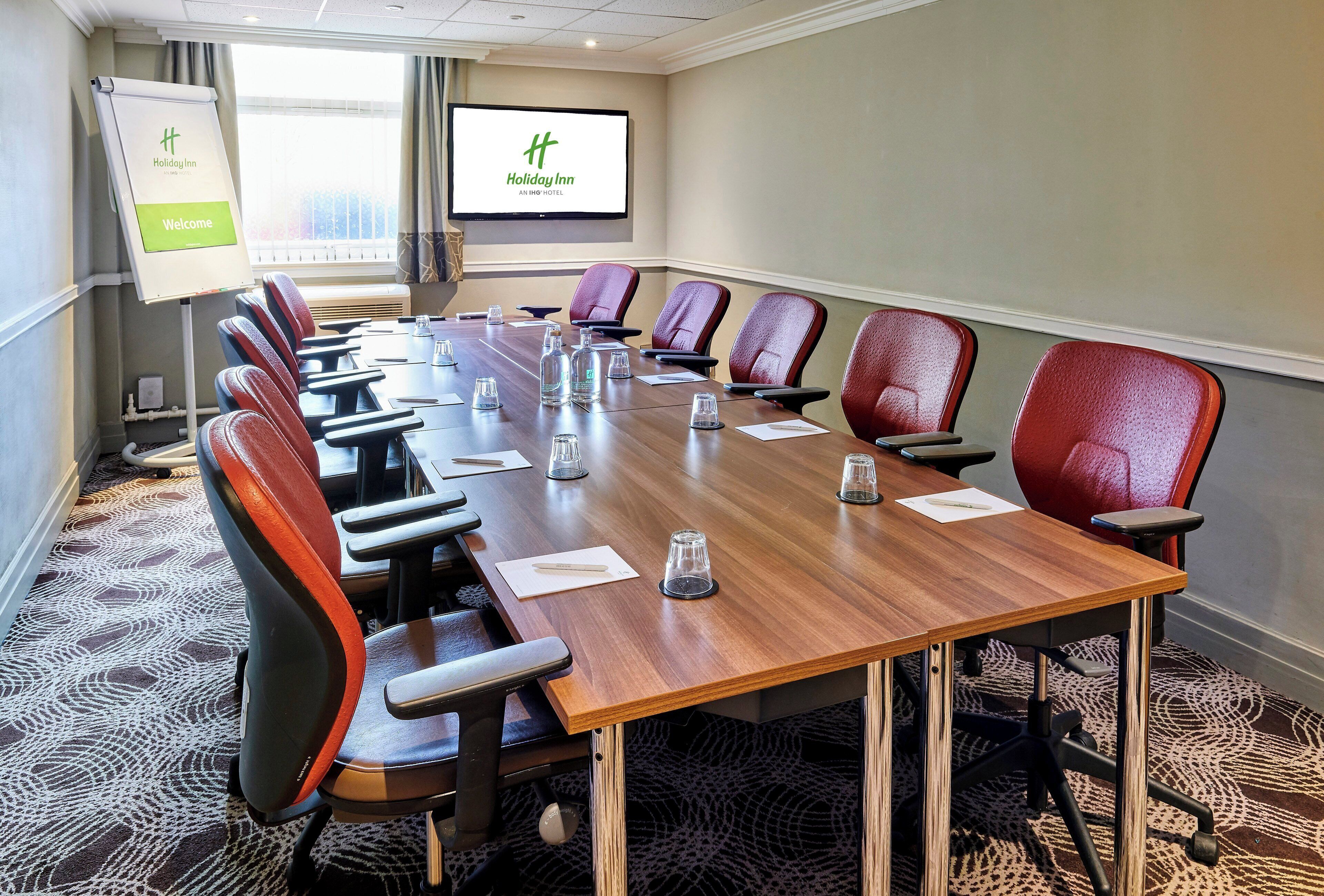 A conference room with a long table, red chairs, a whiteboard, and a screen displaying the Holiday Inn logo.