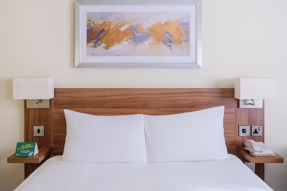 A hotel room with a wooden headboard, white bedding, a framed abstract painting, bedside lamps, and a telephone on a wooden nightstand.