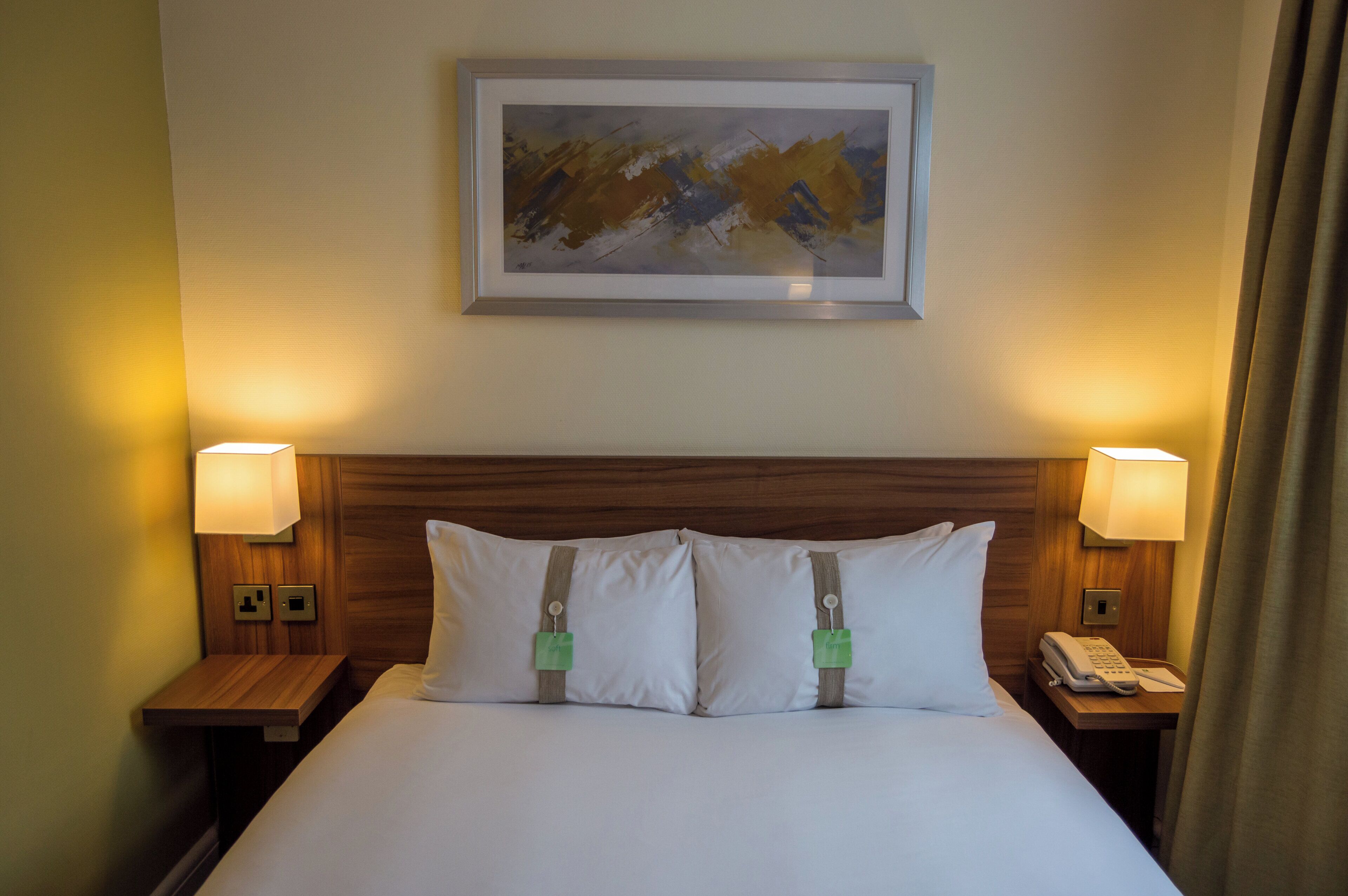 Una camera d'albergo con un letto grande, due lampade sui comodini, un quadro appeso al muro e un telefono sul tavolino.