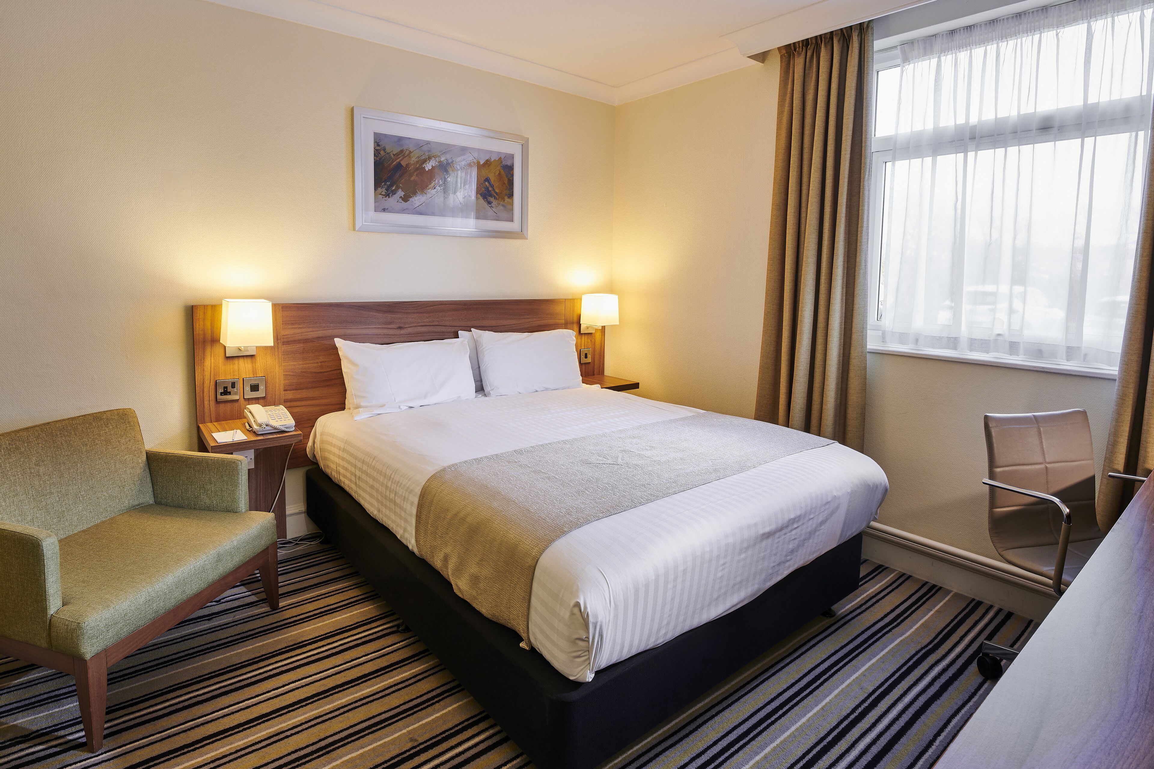 A hotel room with a large bed, two bedside lamps, a chair, a desk with a phone, and a window with curtains.