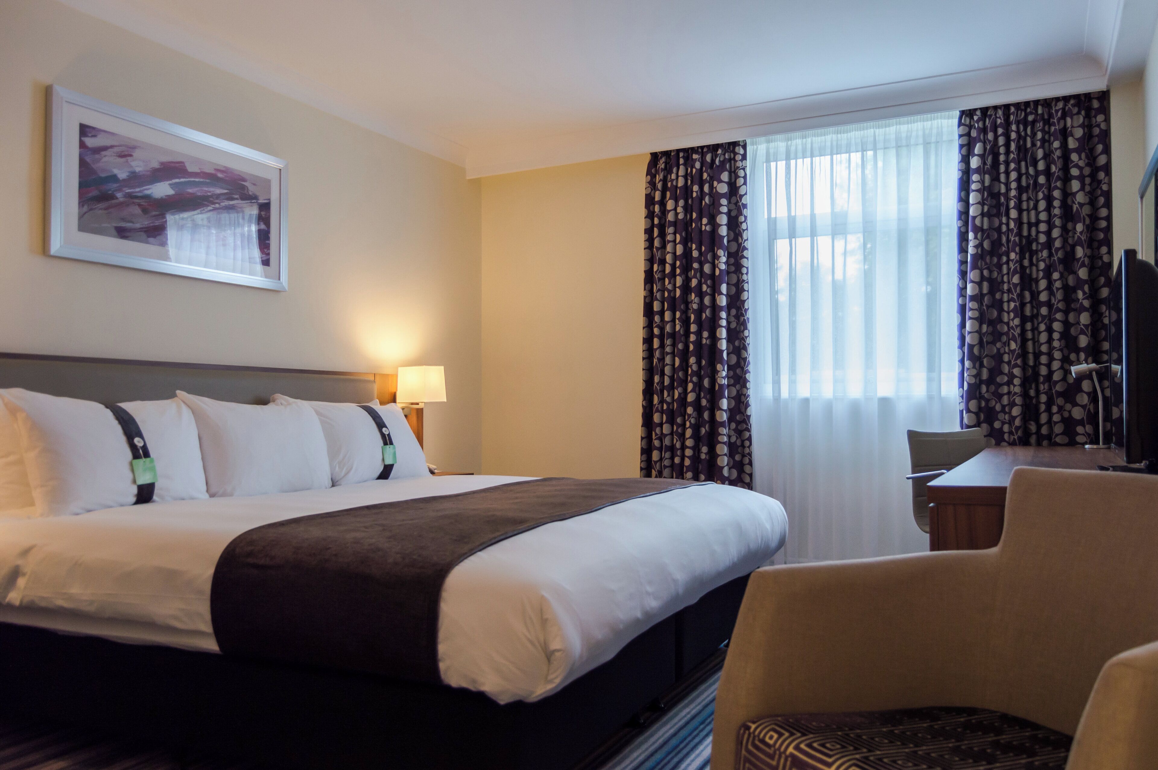 A hotel room with a bed, a chair, a desk, a lamp, and curtains.