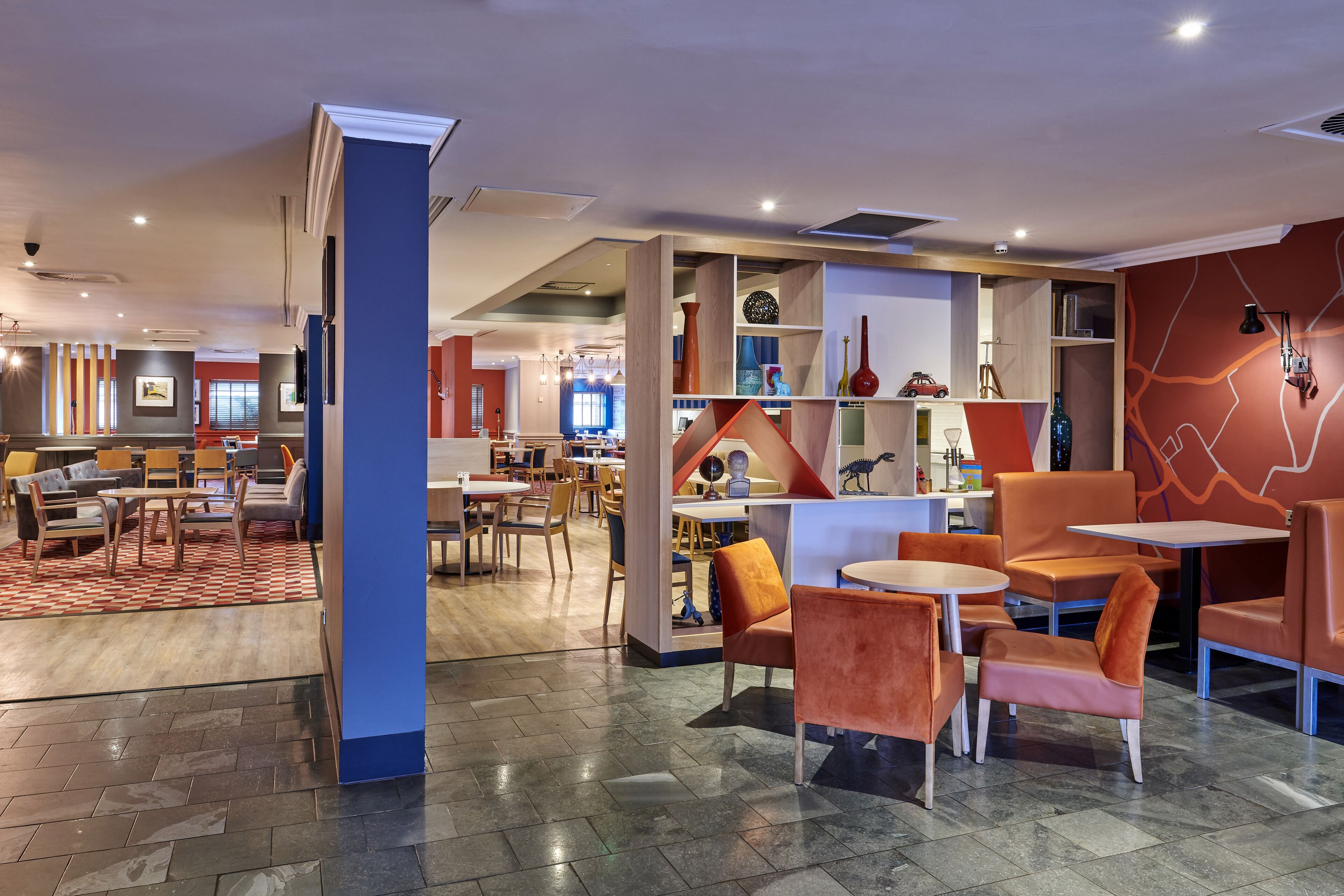 Un moderno vestíbulo de hotel con diversas zonas de estar, incluidas mesas y sillas, y un espacio central con un pilar azul y una estantería.