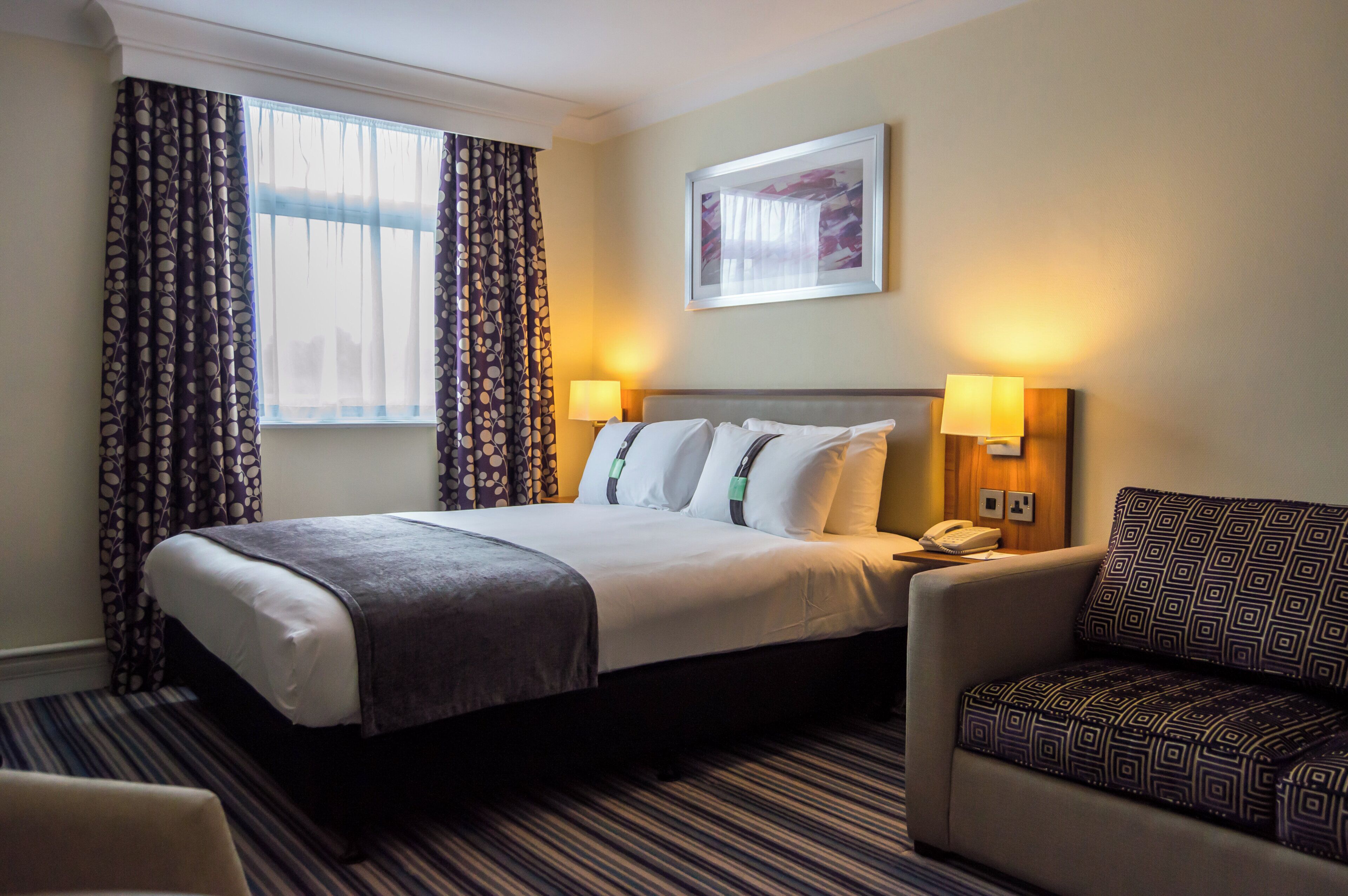 A hotel room with a bed, two bedside lamps, a chair, a window with curtains, and a painting on the wall.