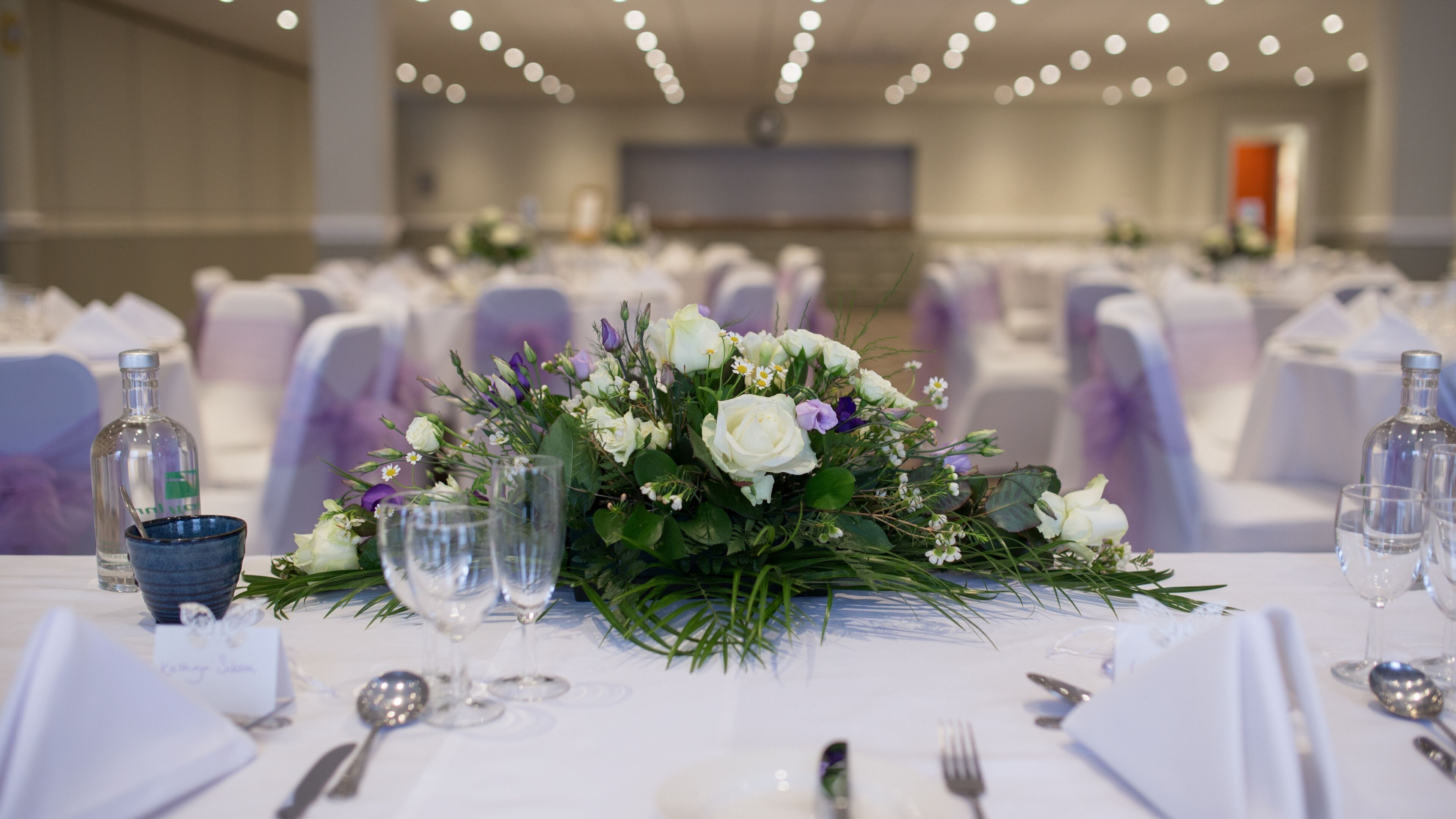 Una mesa de banquete dispuesta con manteles blancos, cristalería y un centro de mesa floral.