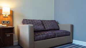 A hotel room with a beige sofa, patterned cushions, a bedside table with a lamp, and a striped carpet. - Holiday Inn Leeds Garforth by IHG (Leeds)
