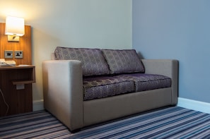 A hotel room with a beige sofa, patterned cushions, a bedside table with a lamp, and a striped carpet.