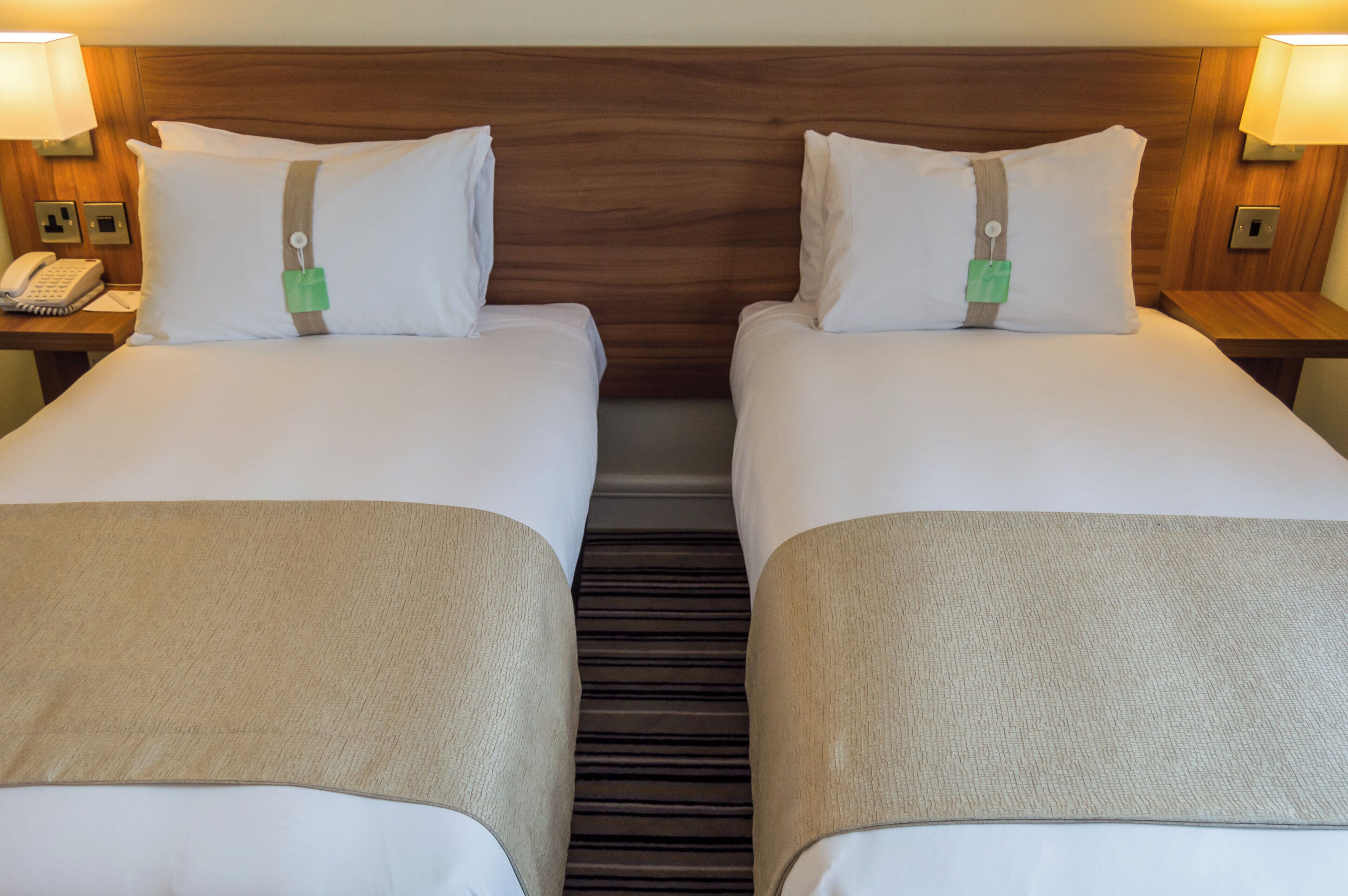 A hotel room with two beds, each with a pillow and a tag, a bedside phone, and a lamp on the wall.