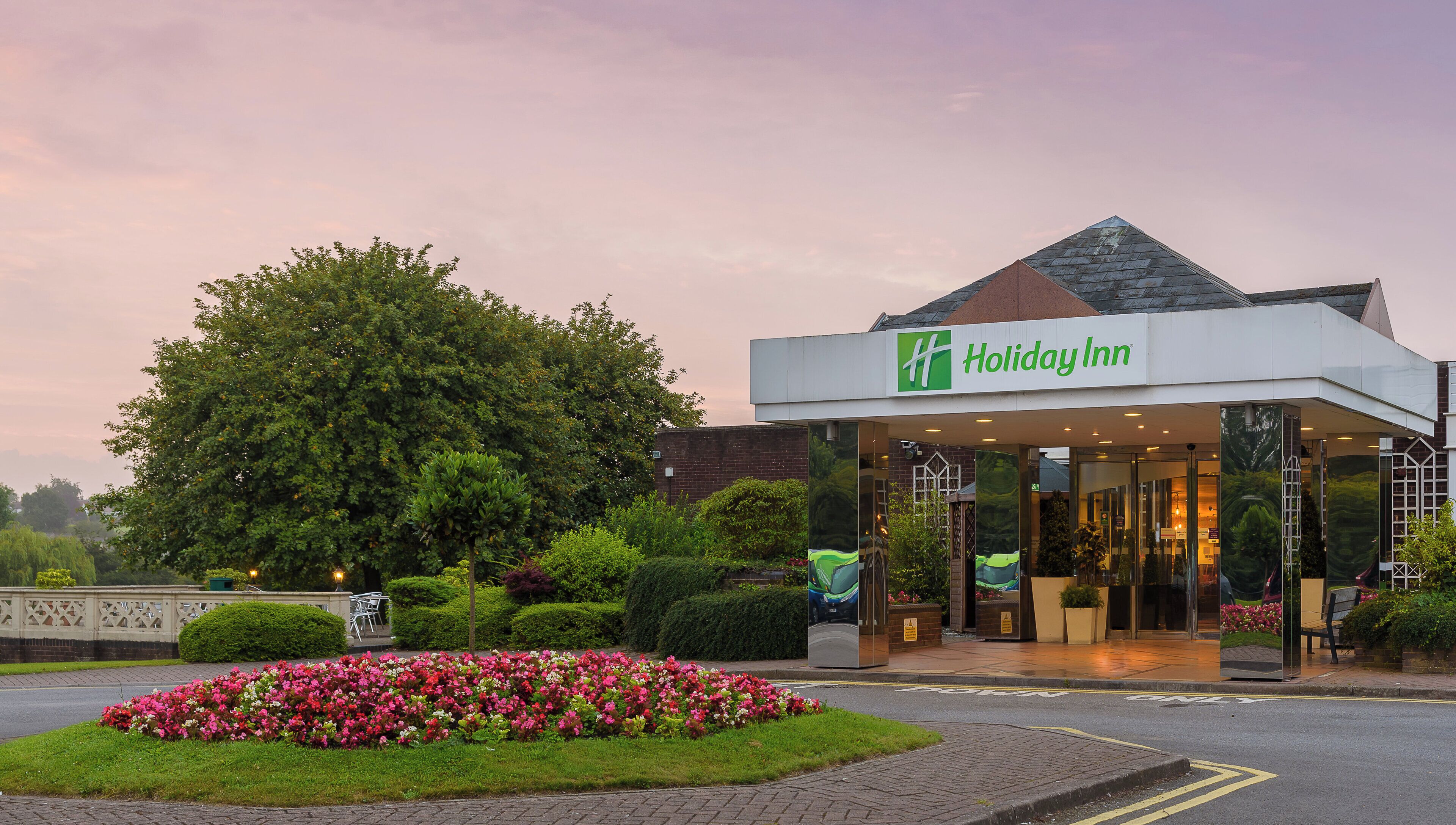 A Holiday Inn hotel entrance with a modern design, a landscaped garden, and a large tree.