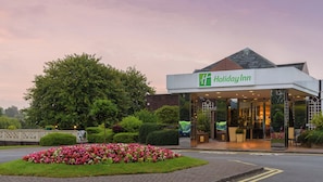 Entrada de un hotel Holiday Inn con un diseño moderno, un jardín ajardinado y un árbol grande.