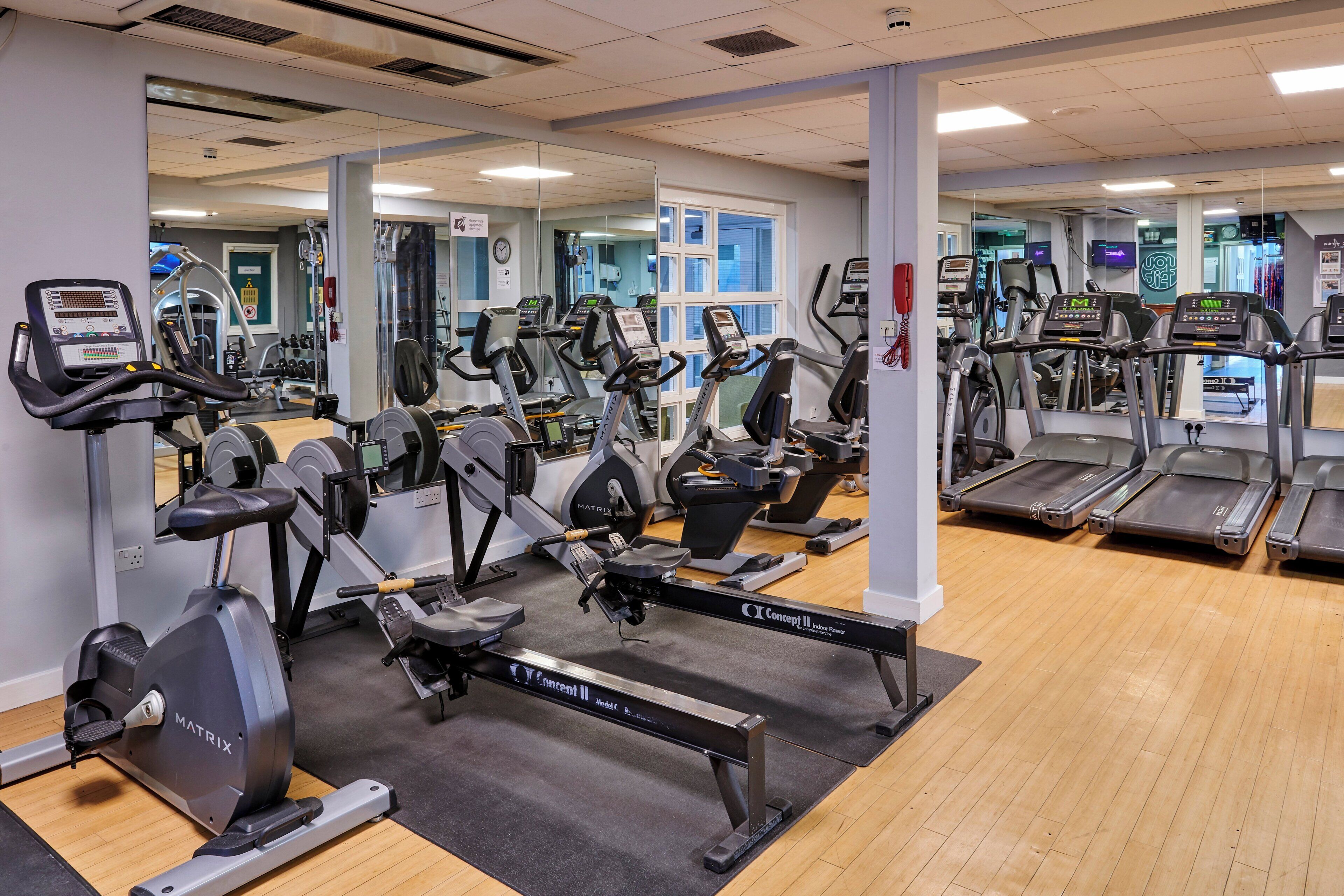 A gym with multiple cardio machines, mirrors, and a wooden floor.
