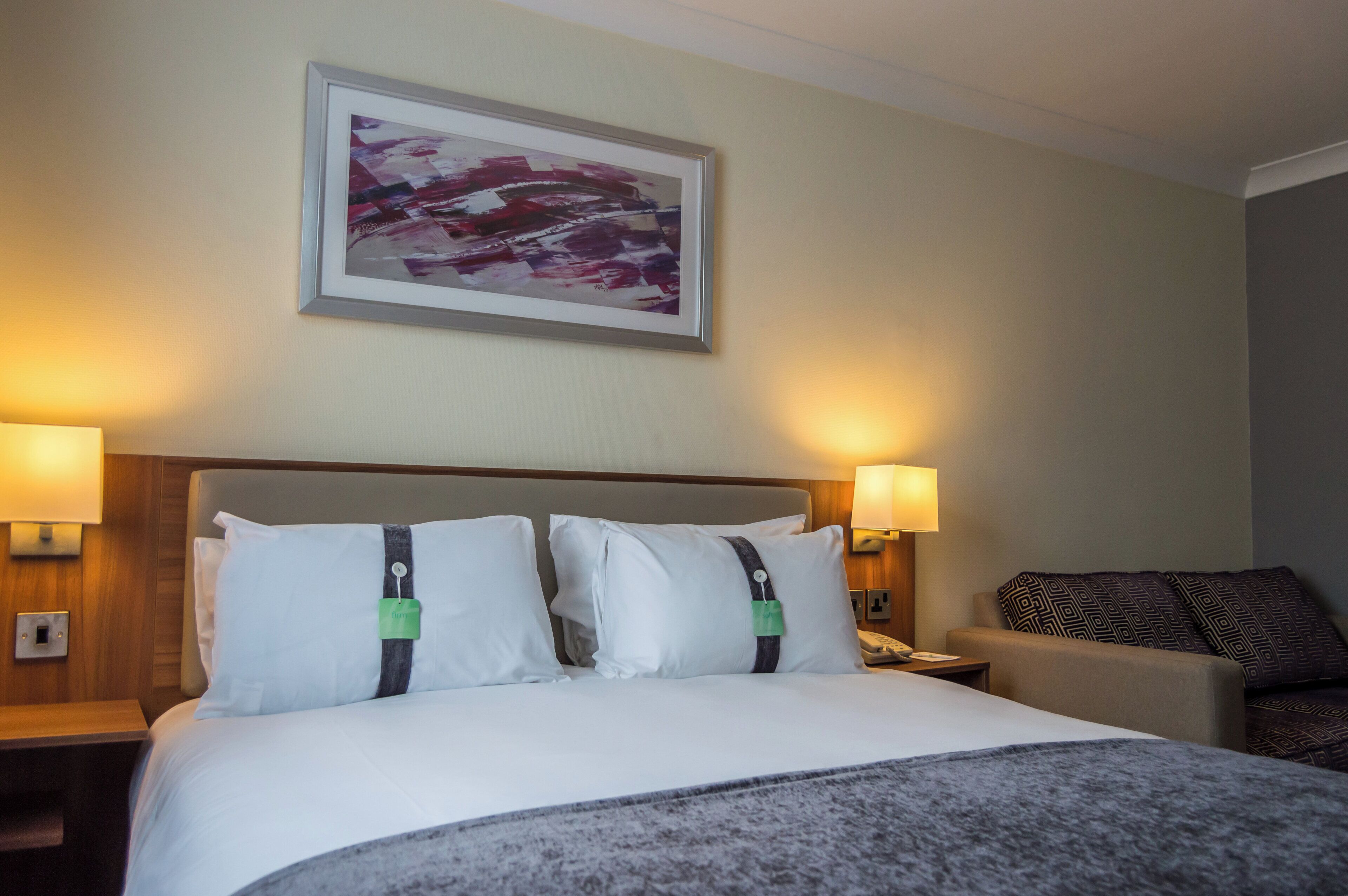 Una habitación de hotel con una cama grande, dos lámparas de noche, un cuadro abstracto enmarcado y un plaid estampado sobre la cama.
