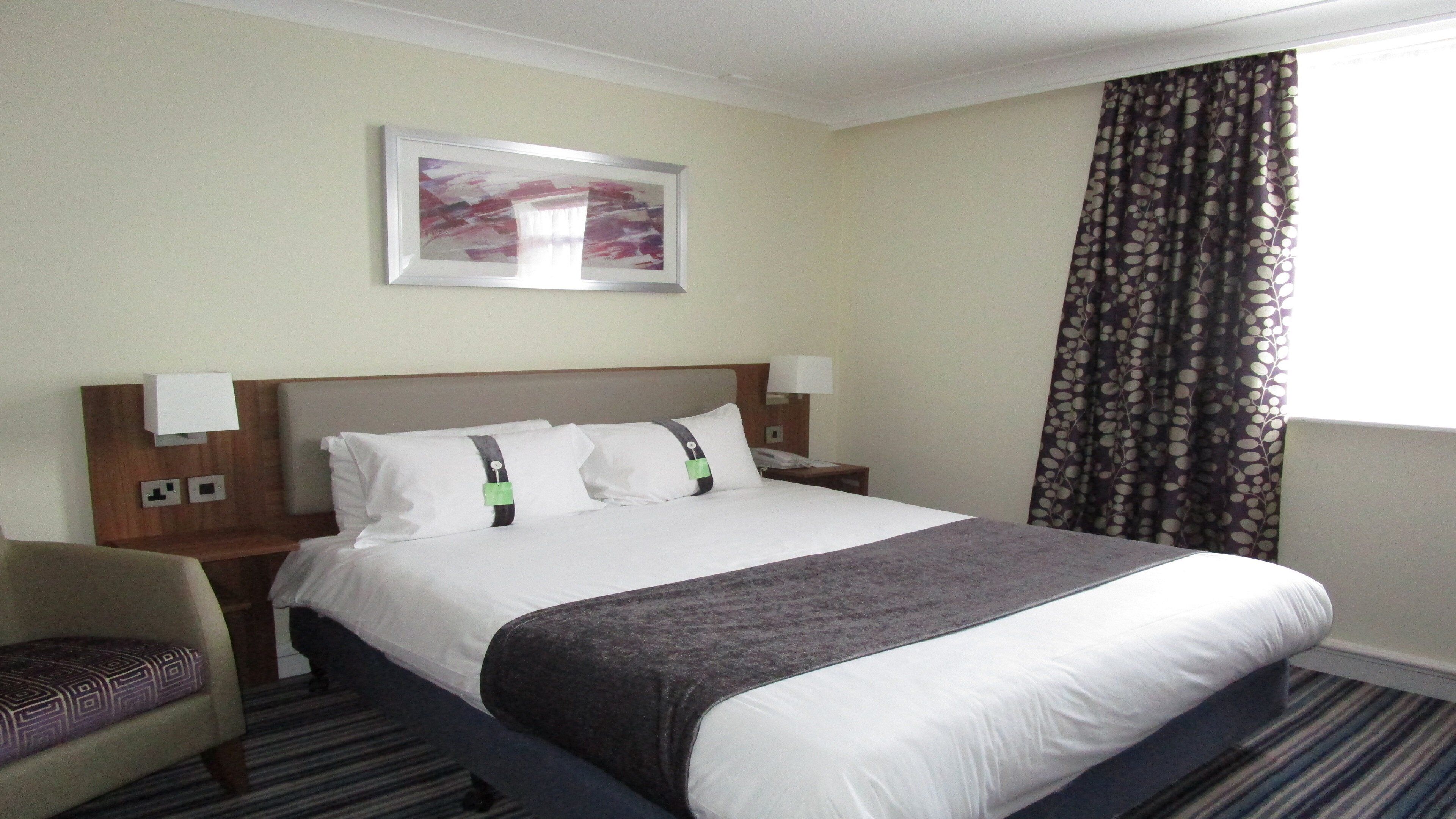 A hotel room with a bed, a nightstand, a chair, a painting on the wall, and a window with curtains.