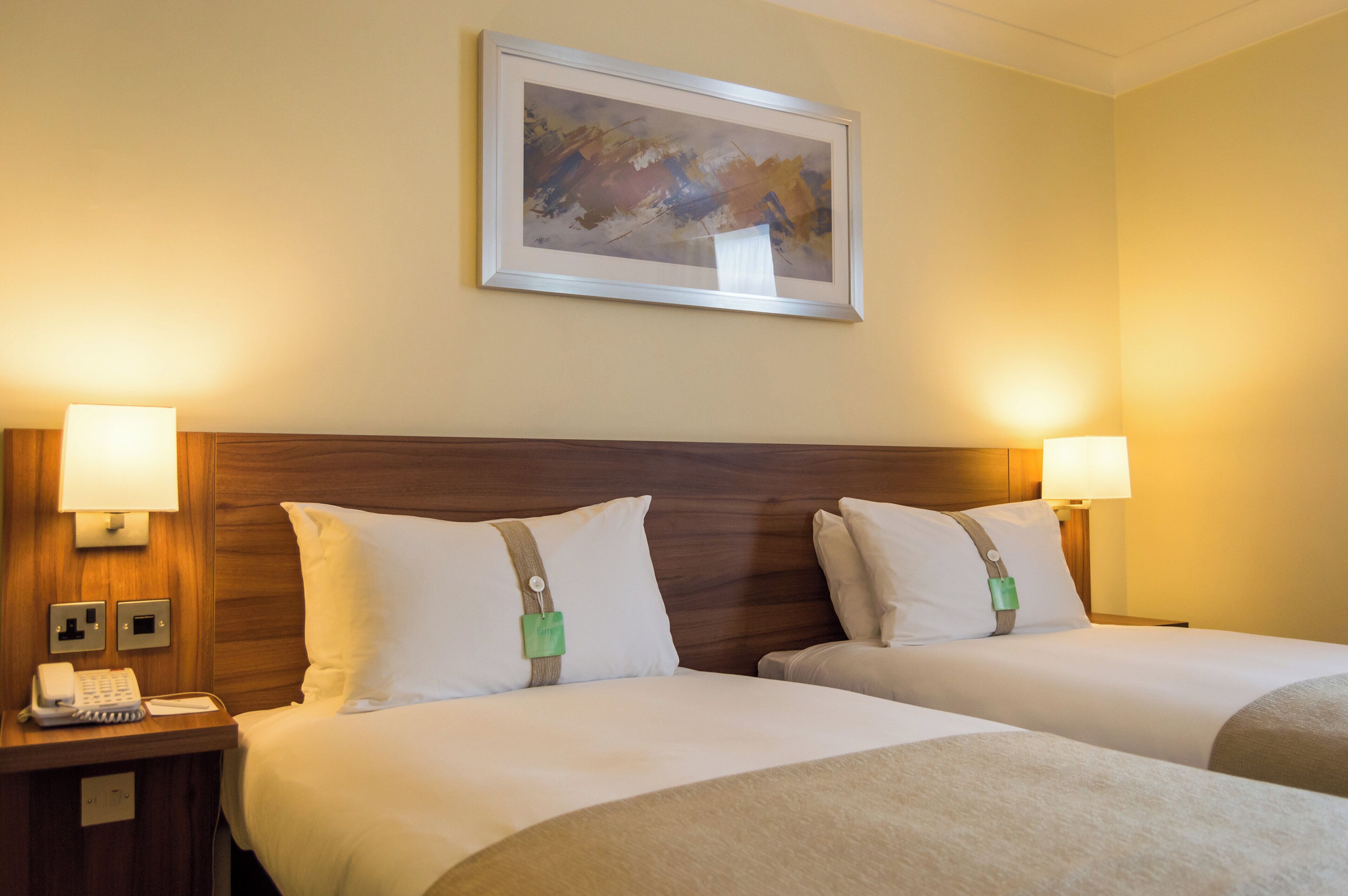 A hotel room with two beds, a wooden headboard, bedside lamps, a telephone, and a painting on the wall.