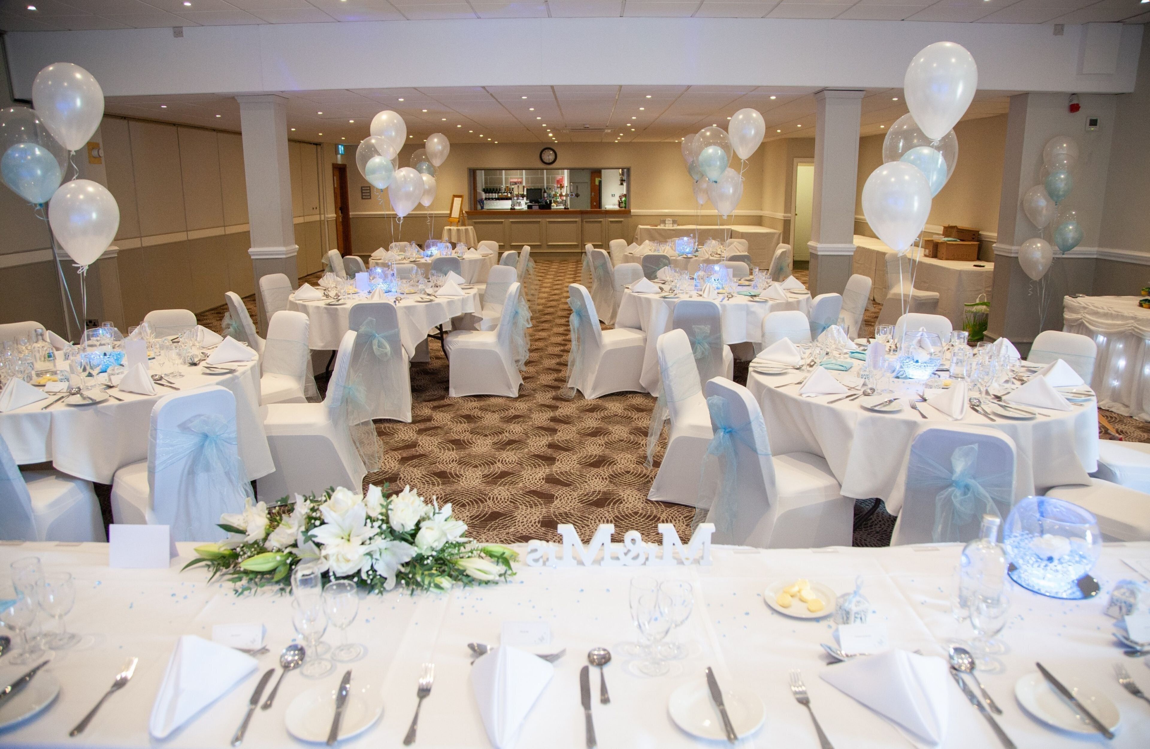 A banquet hall with round tables set for a formal event, featuring white tablecloths, chairs with sashes, and a carpeted floor.