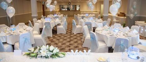 A banquet hall with round tables set for a formal event, featuring white tablecloths, chairs with sashes, and a carpeted floor.