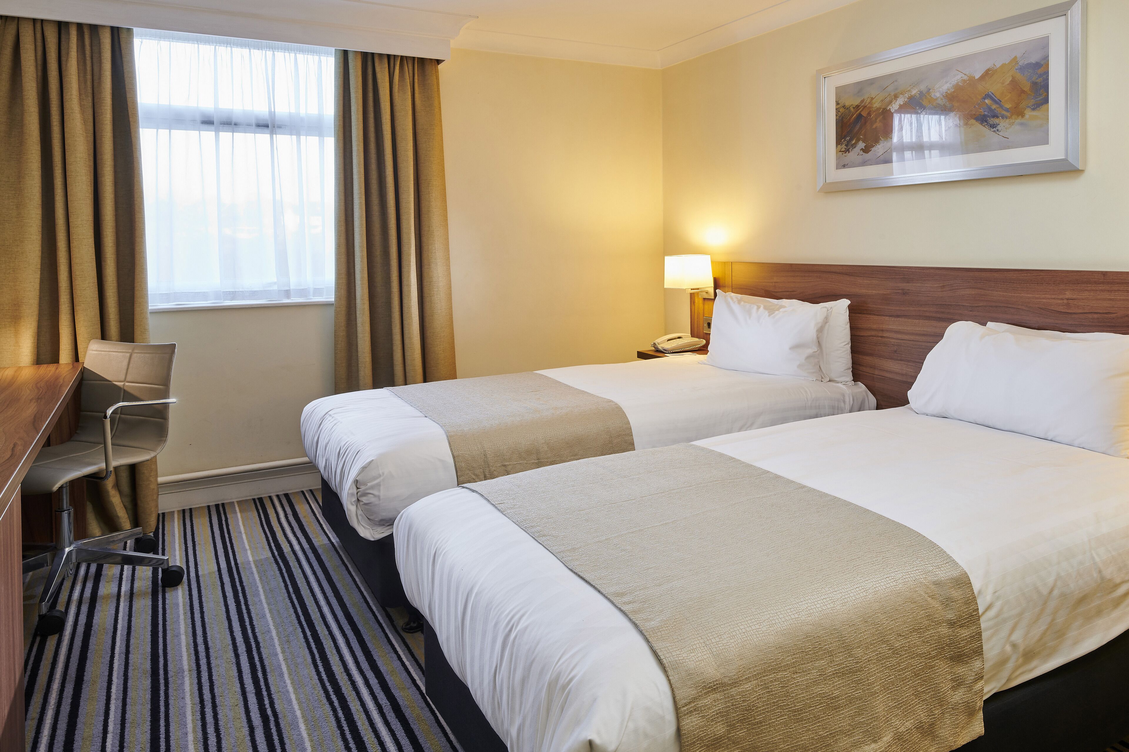 A hotel room with two beds, a desk, a chair, a lamp, and a painting on the wall.