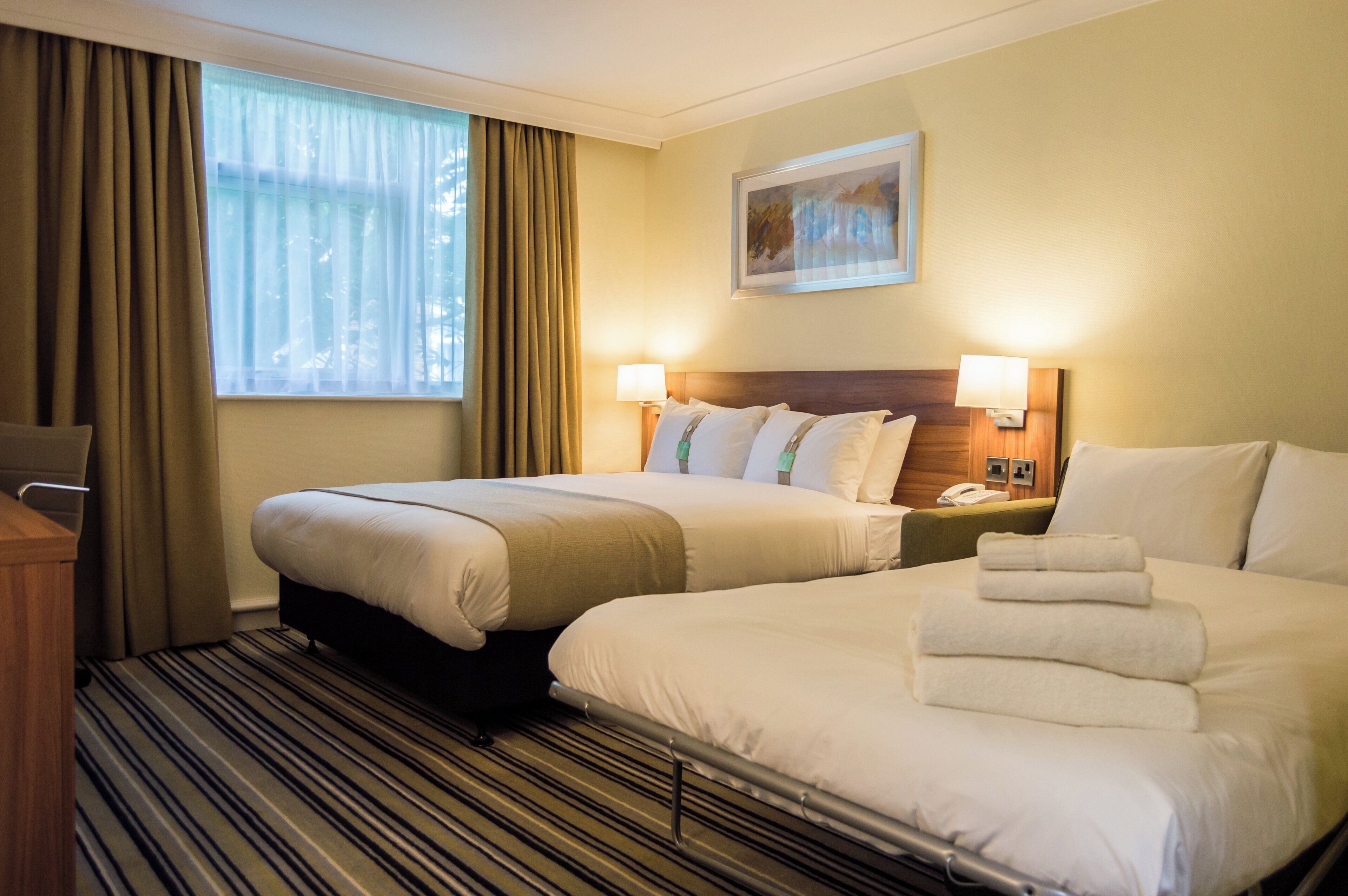 A hotel room with two beds, a window with curtains, a painting on the wall, and a bedside lamp.