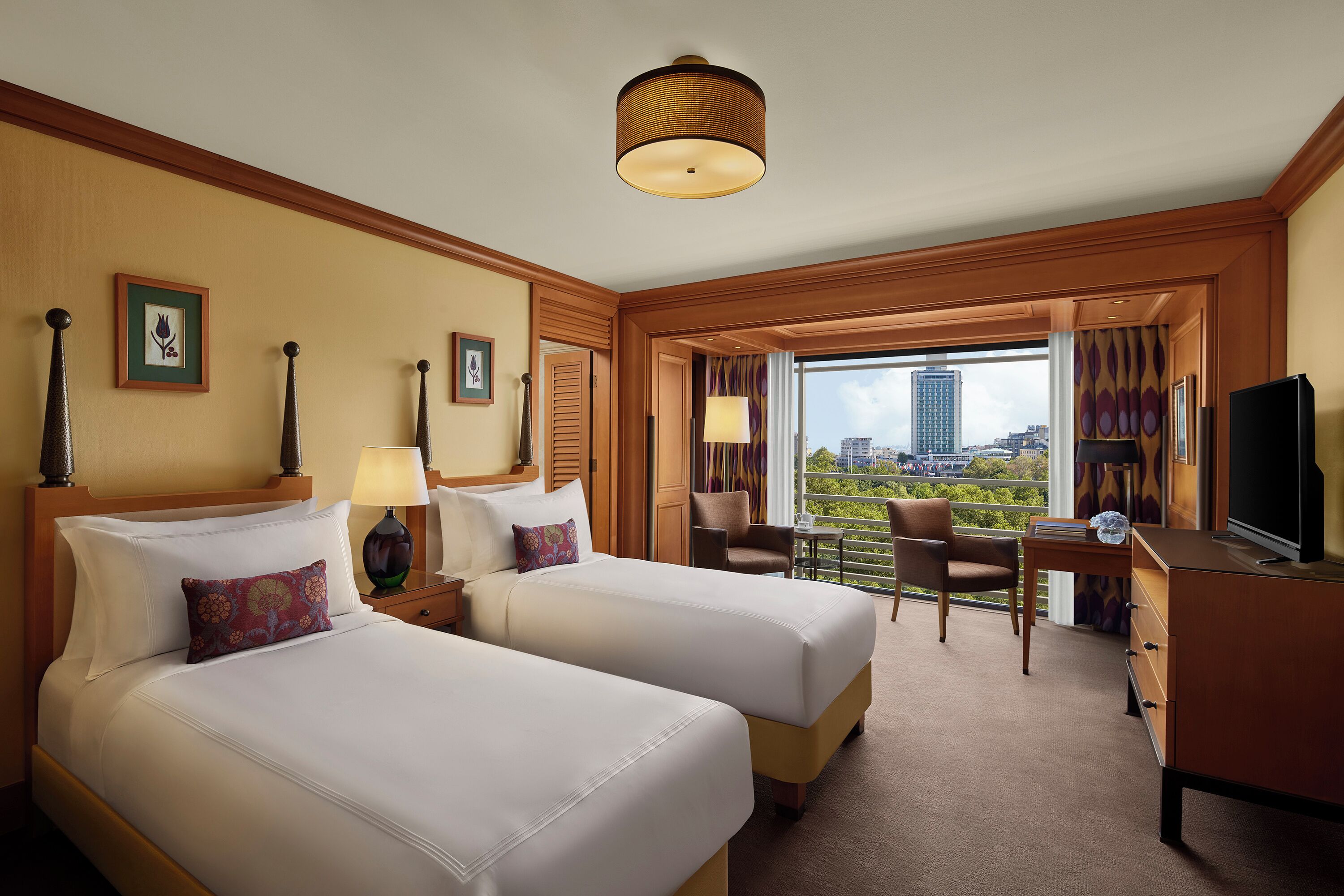 A hotel room with two beds, a desk, a chair, a television, and a view of the city.