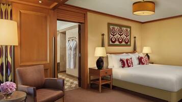 A hotel room with a bed, two bedside lamps, a chair, a small table with a vase, and a door leading to a bathroom.
