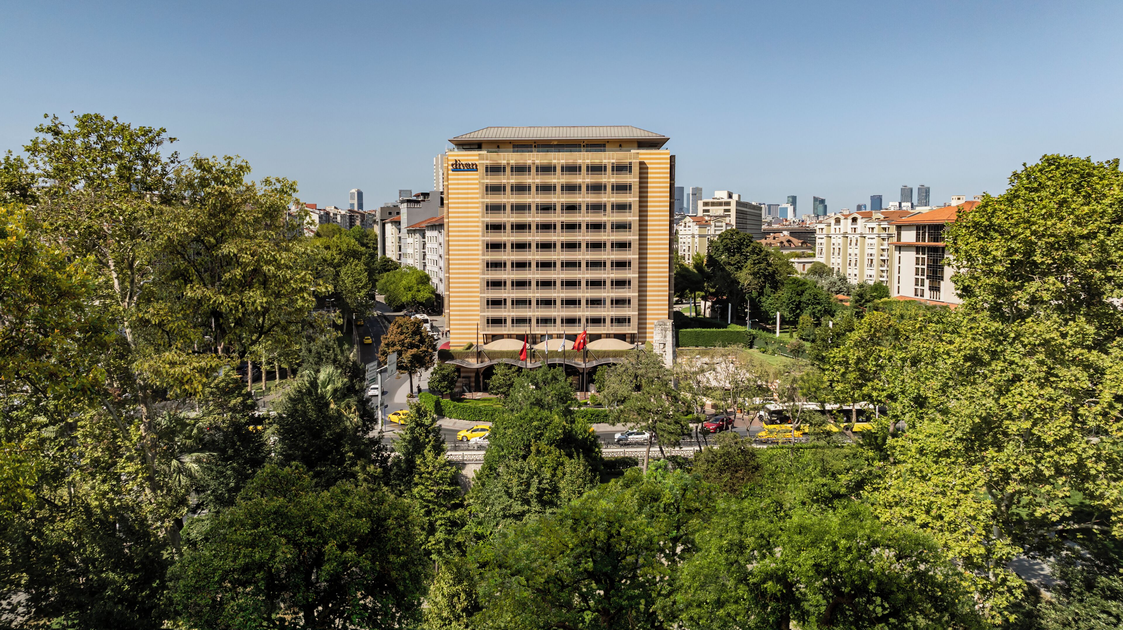 A high-rise building with a grid-like facade, surrounded by dense greenery and other buildings in the background.