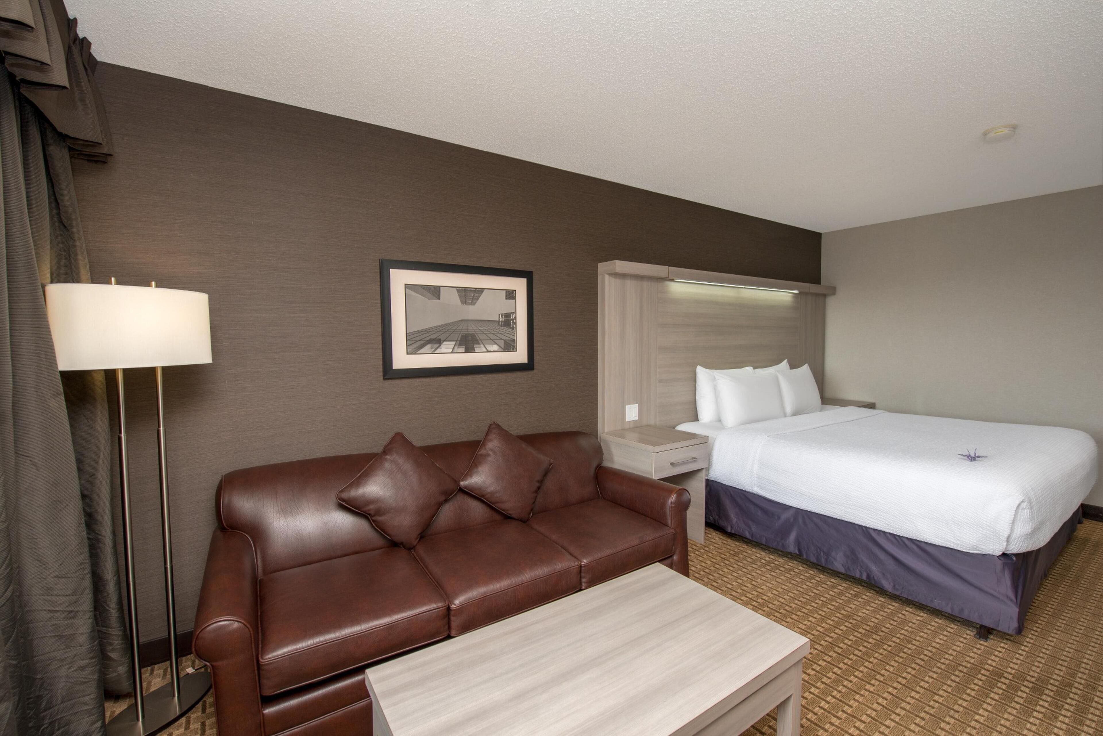 A hotel room with a brown leather sofa, a small table, a bed with white linens, a nightstand, a lamp, and a framed picture on the wall.