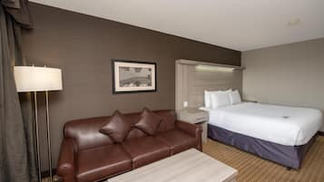 A hotel room with a brown leather sofa, a small table, a bed with white linens, a nightstand, a lamp, and a framed picture on the wall.