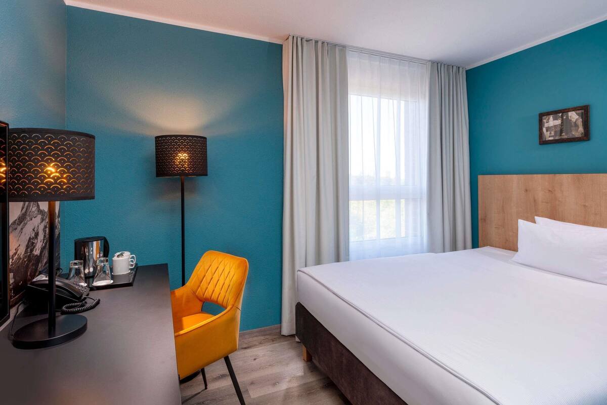 A hotel room with a bed, a desk with a lamp, a chair, and a window with curtains.