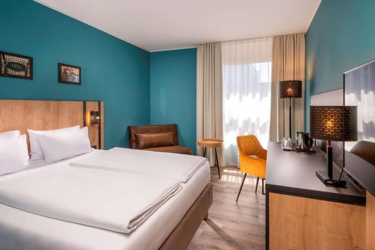 A hotel room with a large bed, a desk with a lamp, a chair, and a city view.
